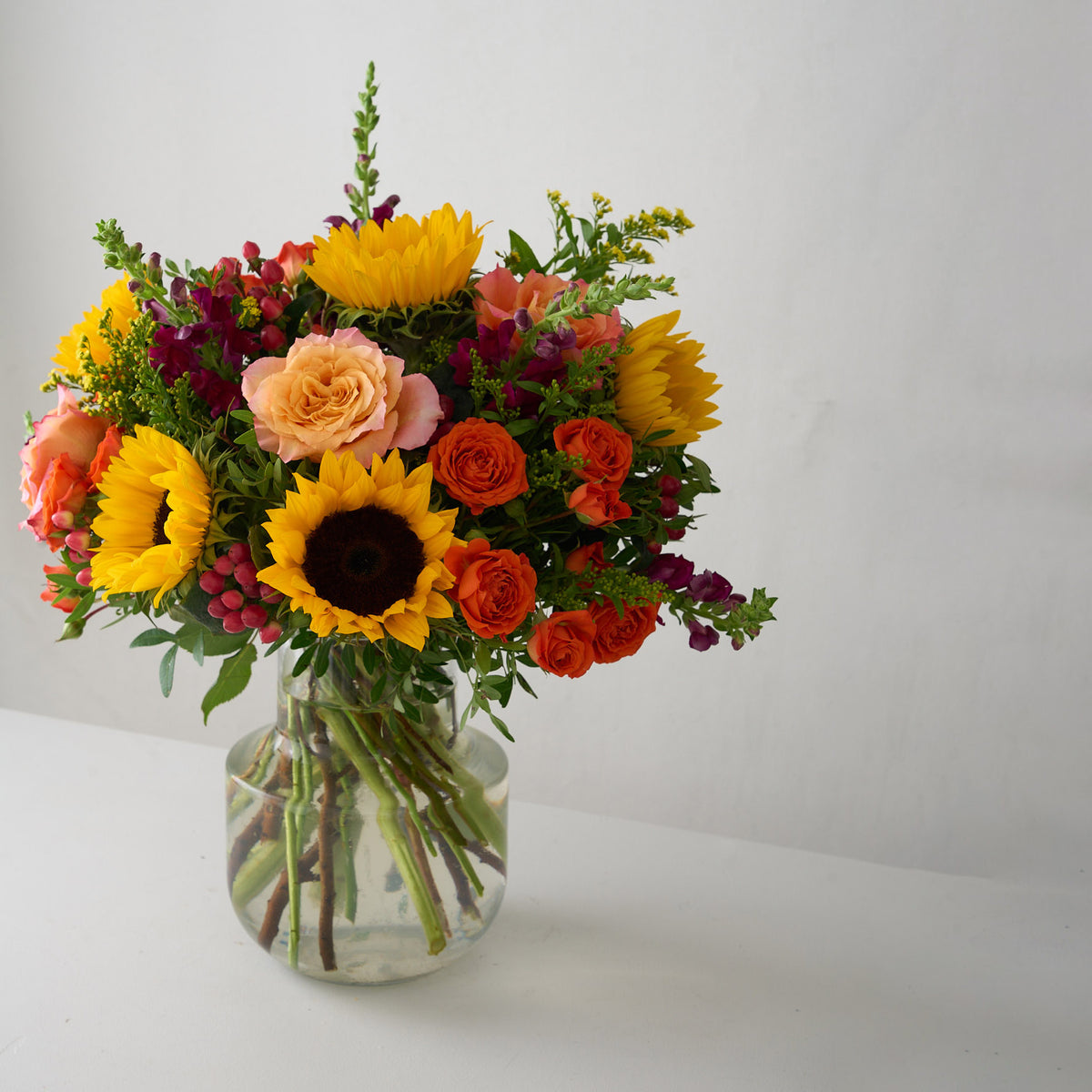 A vibrant autumn flower bouquet in a clear glass vase, featuring yellow sunflowers, orange and pink roses, and purple berries on a white surface against a light gray wall.