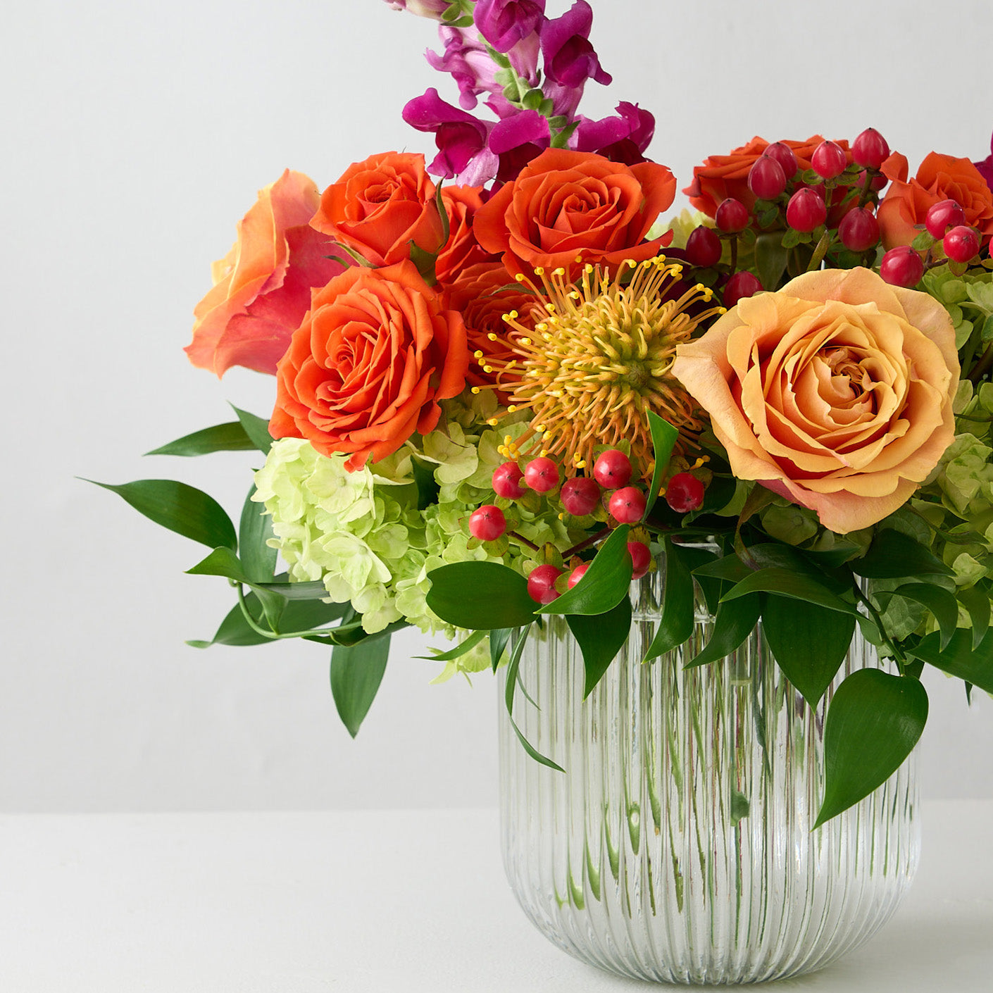 zoomed in picture of coral roses and hypericum, green hydrangea , orange spray roses, fuchsia snapdragons, and protea in a clear ribbed short cylinder vase