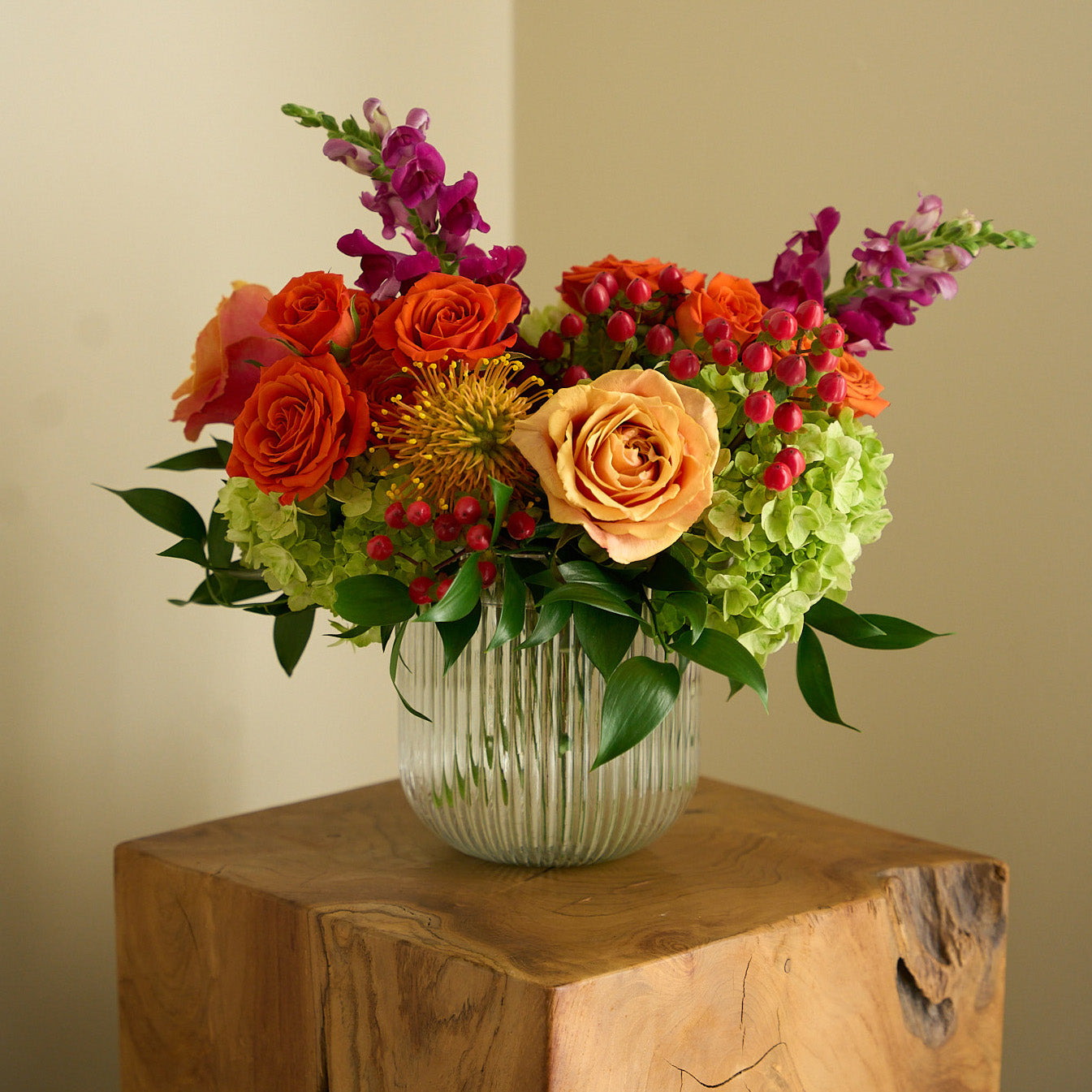 coral roses and hypericum, green hydrangea , orange spray roses, fuchsia snapdragons, and protea in a clear ribbed short cylinder vase on a wooden side table