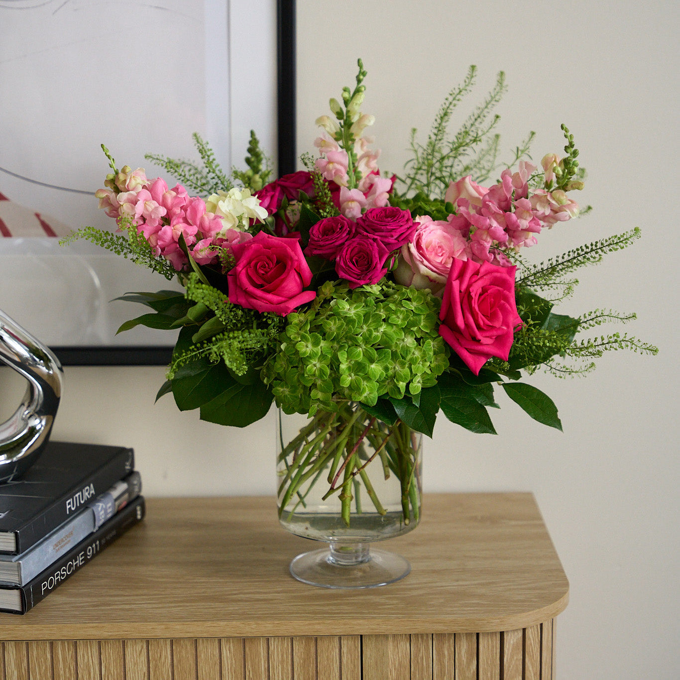 valentine's day arrangement with pink, hot pick roses, snap dragons, and hydrangea in a clear pedestal vase, on a modern credenza with books