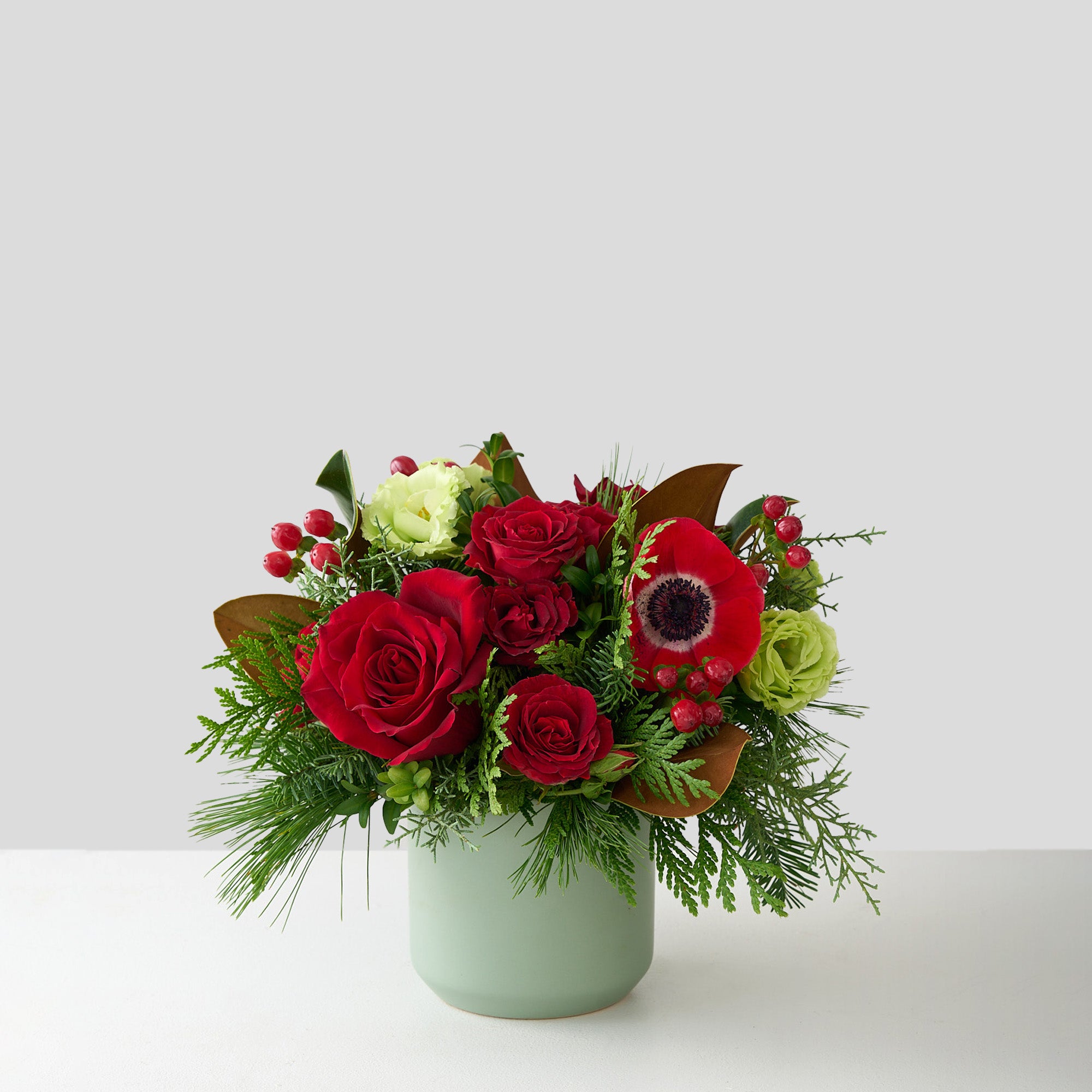Christmas arrangement of red roses, hypericum, anemones, magnolia leaves, green Lisianthus and winter greens in a small sage vase