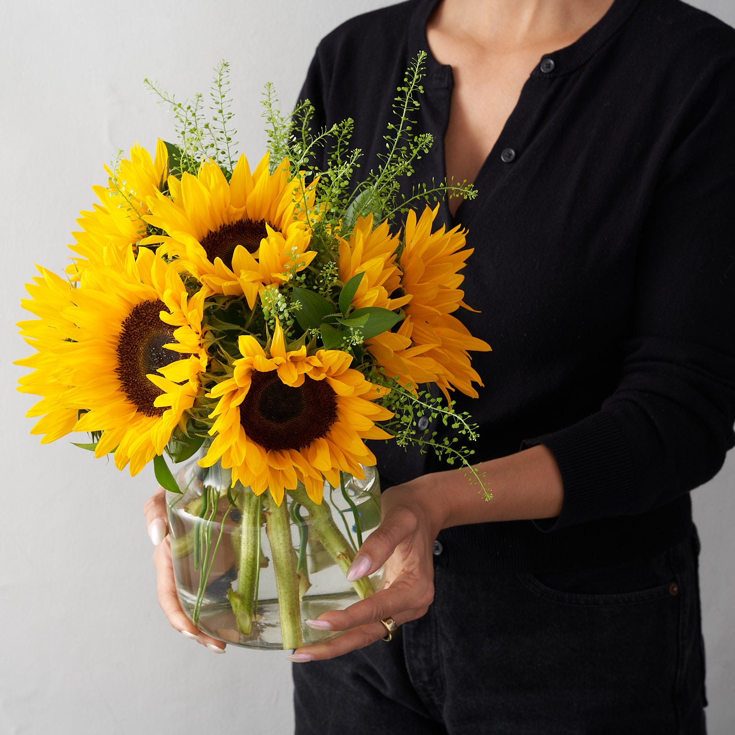 woman in black holding a glass vase of 8 sunflowers with greens