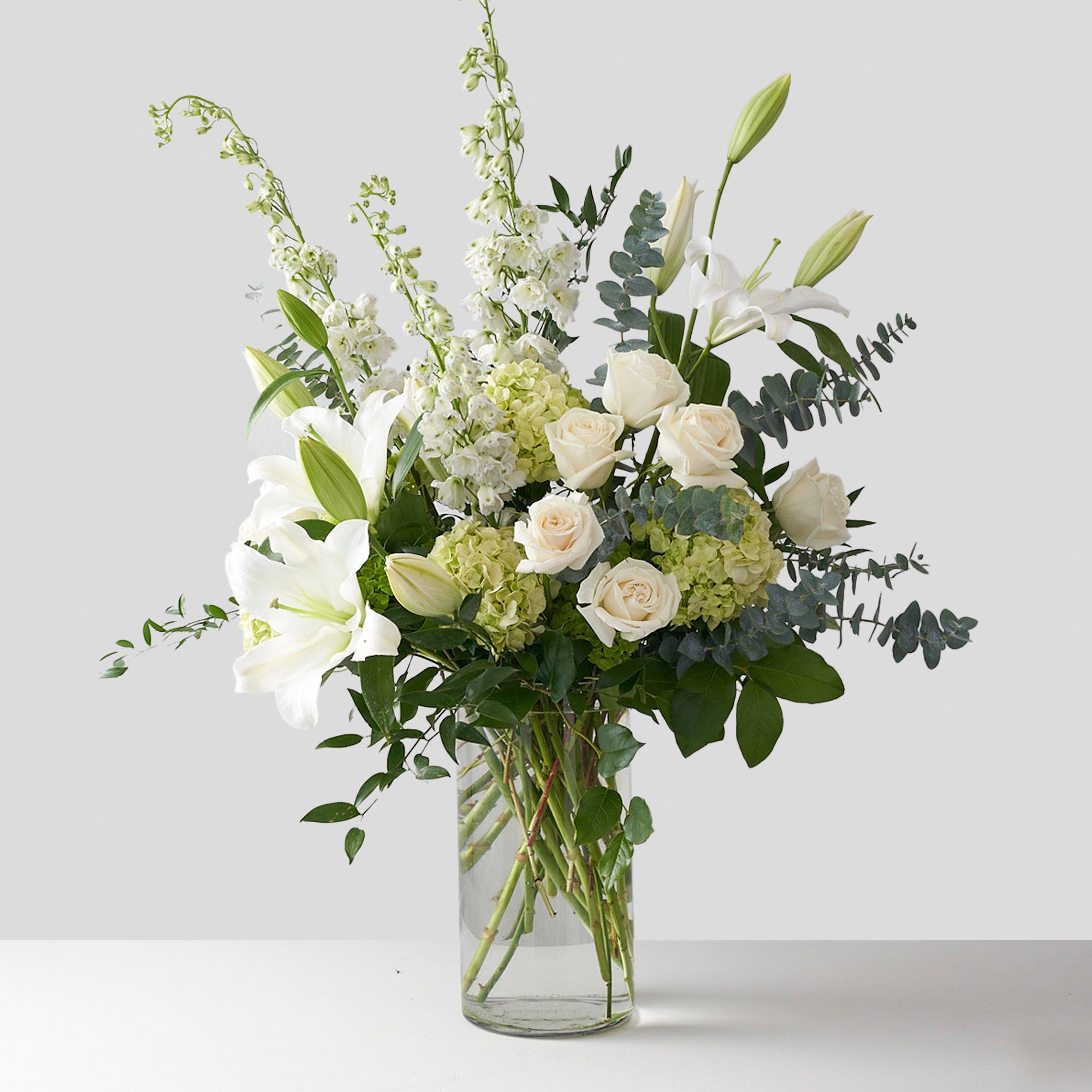 Clear glass vase filled with white and green flowers, featuring white lilies and white roses, green hydrangea, and others