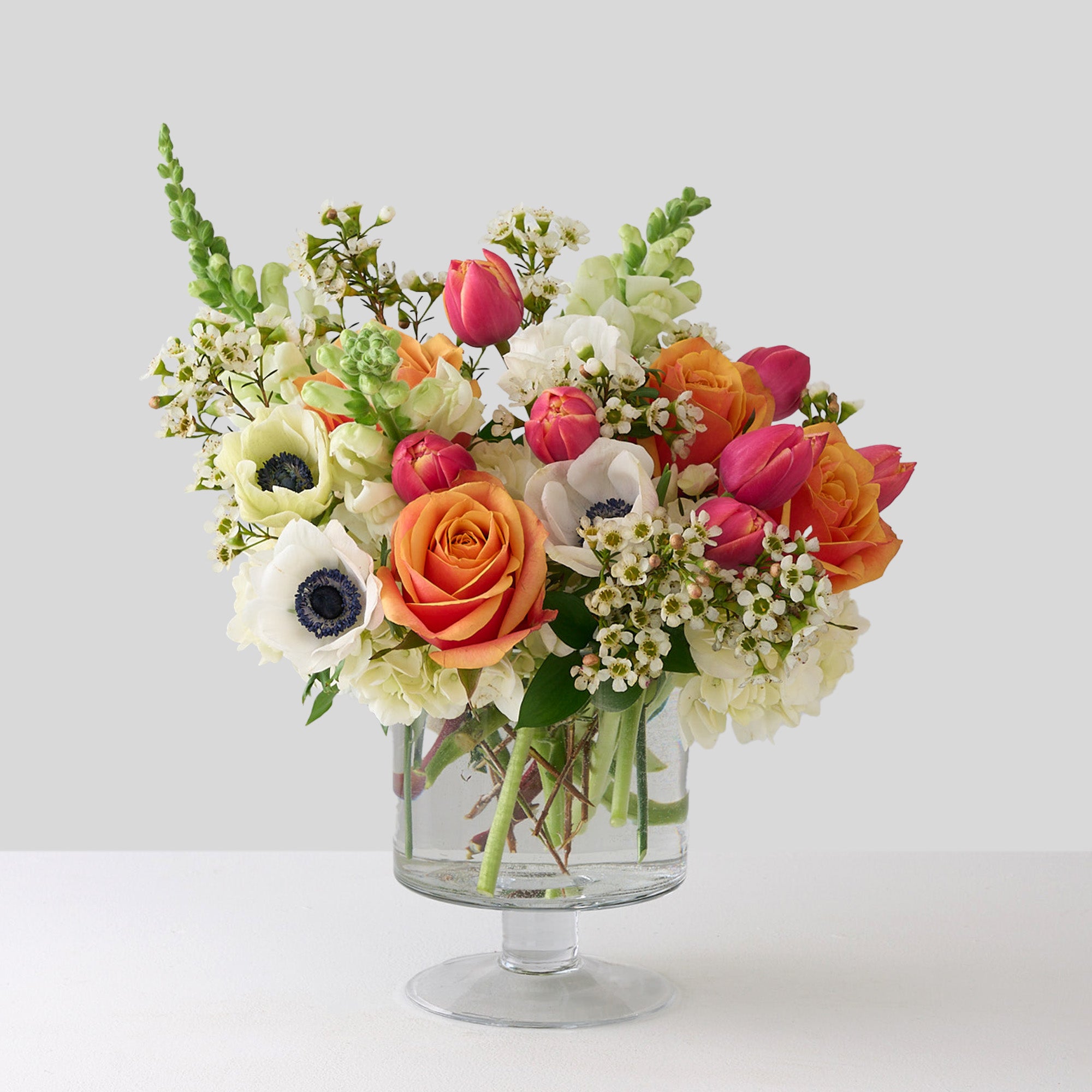 tulips white anemones, snapdragons, wax flowers and orange roses in glass pedestal vase