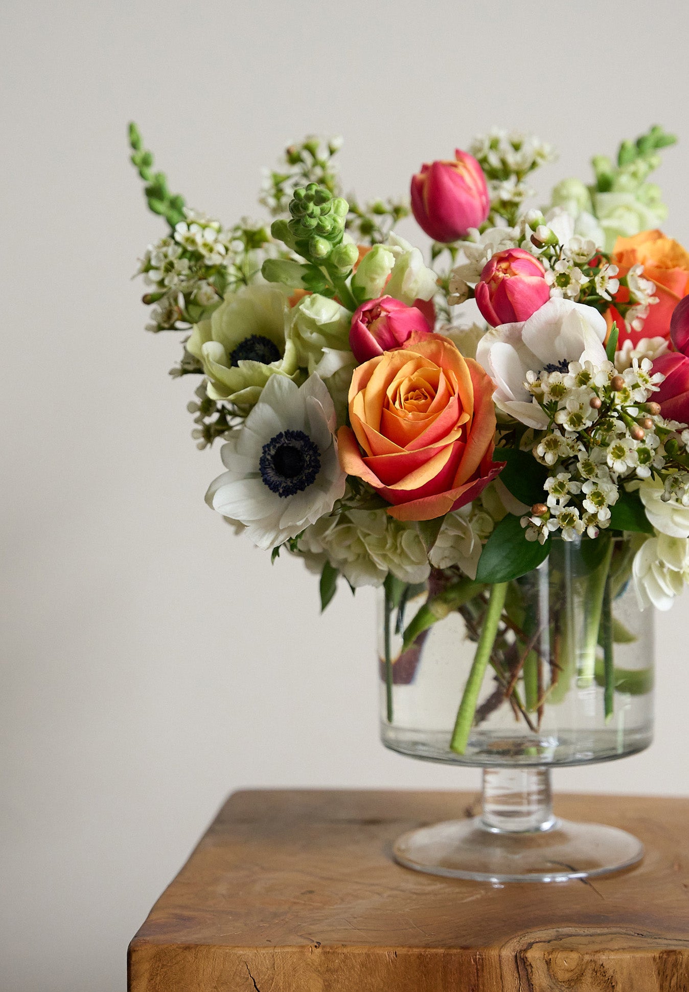 tulips white anemones, snapdragons, wax flowers and orange roses in glass pedestal vase