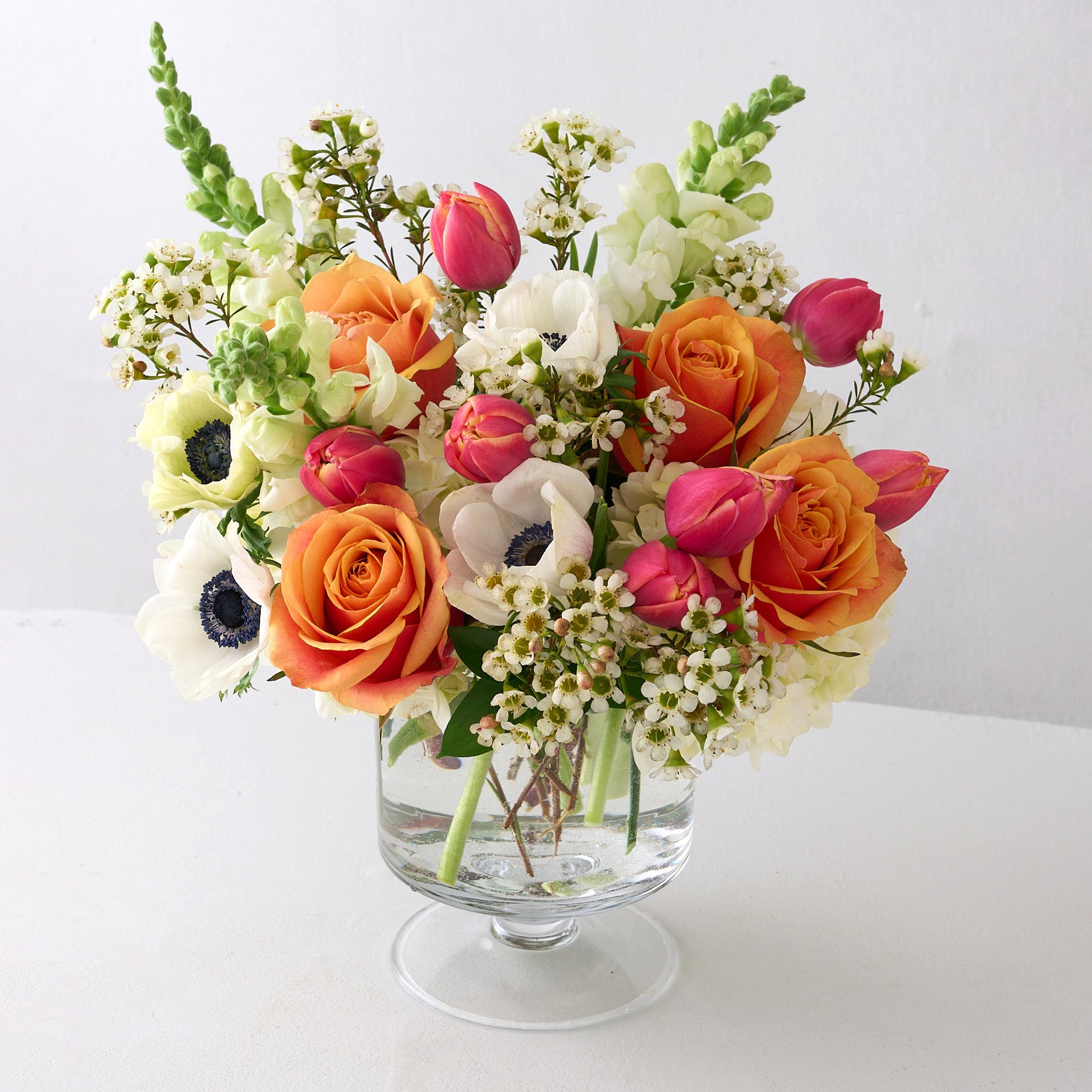 tulips white anemones, snapdragons, wax flowers and orange roses in glass pedestal vase