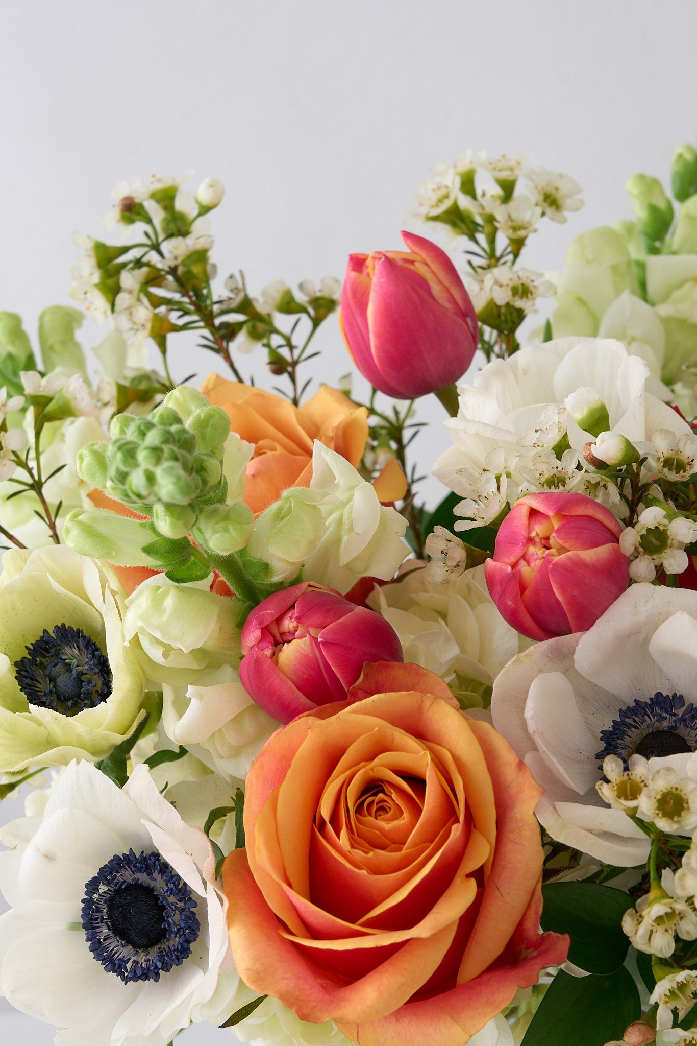 tulips white anemones, snapdragons, wax flowers and orange roses in glass pedestal vase