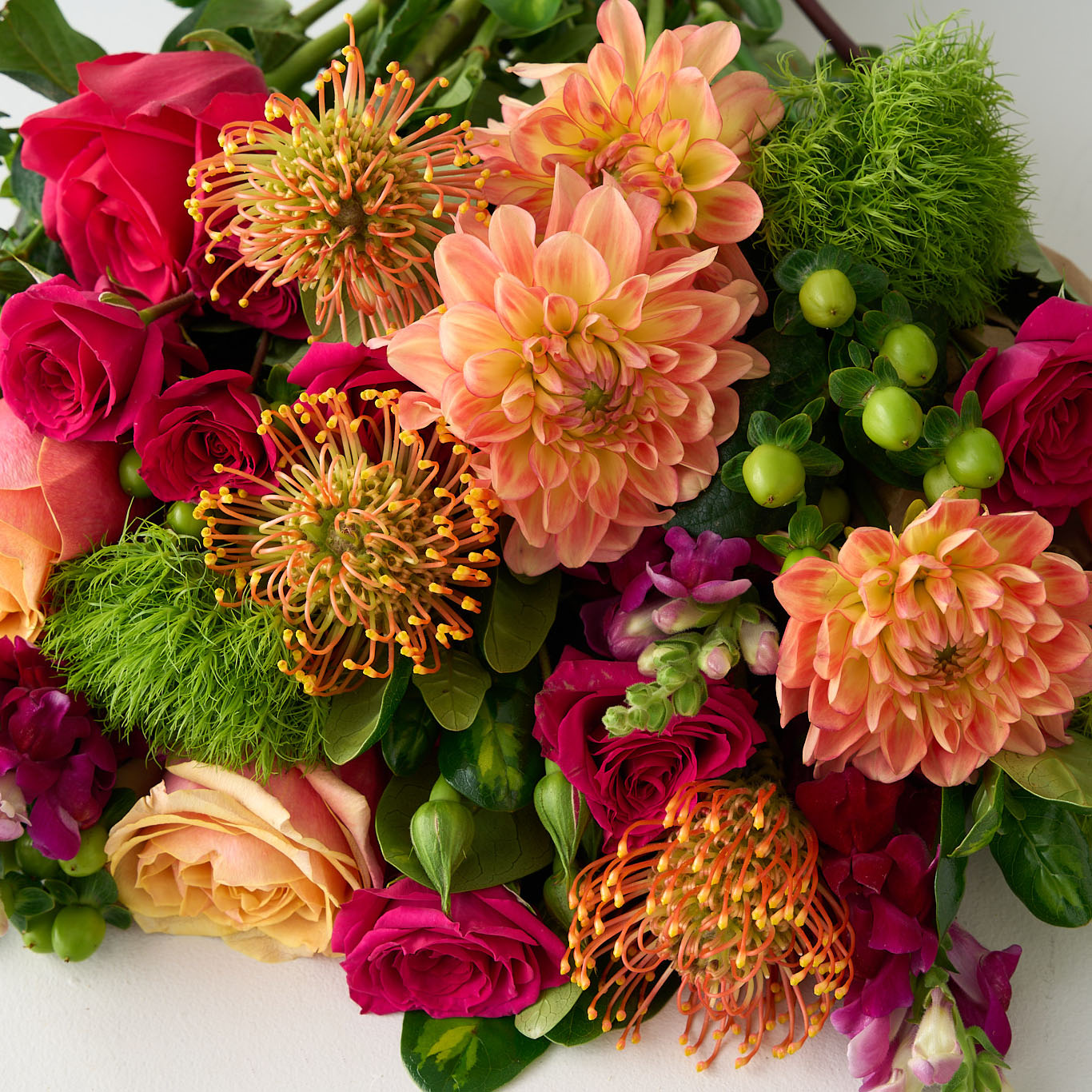 close up photo of a bouquet of colorful flowers including roses, green mums, hypericum and dahlias on a white table