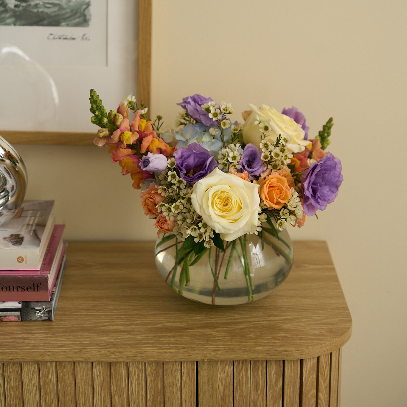 Easter and passover arrangement with peach, cream, blue and purple flowers in a glass vase on a wooden credenza.