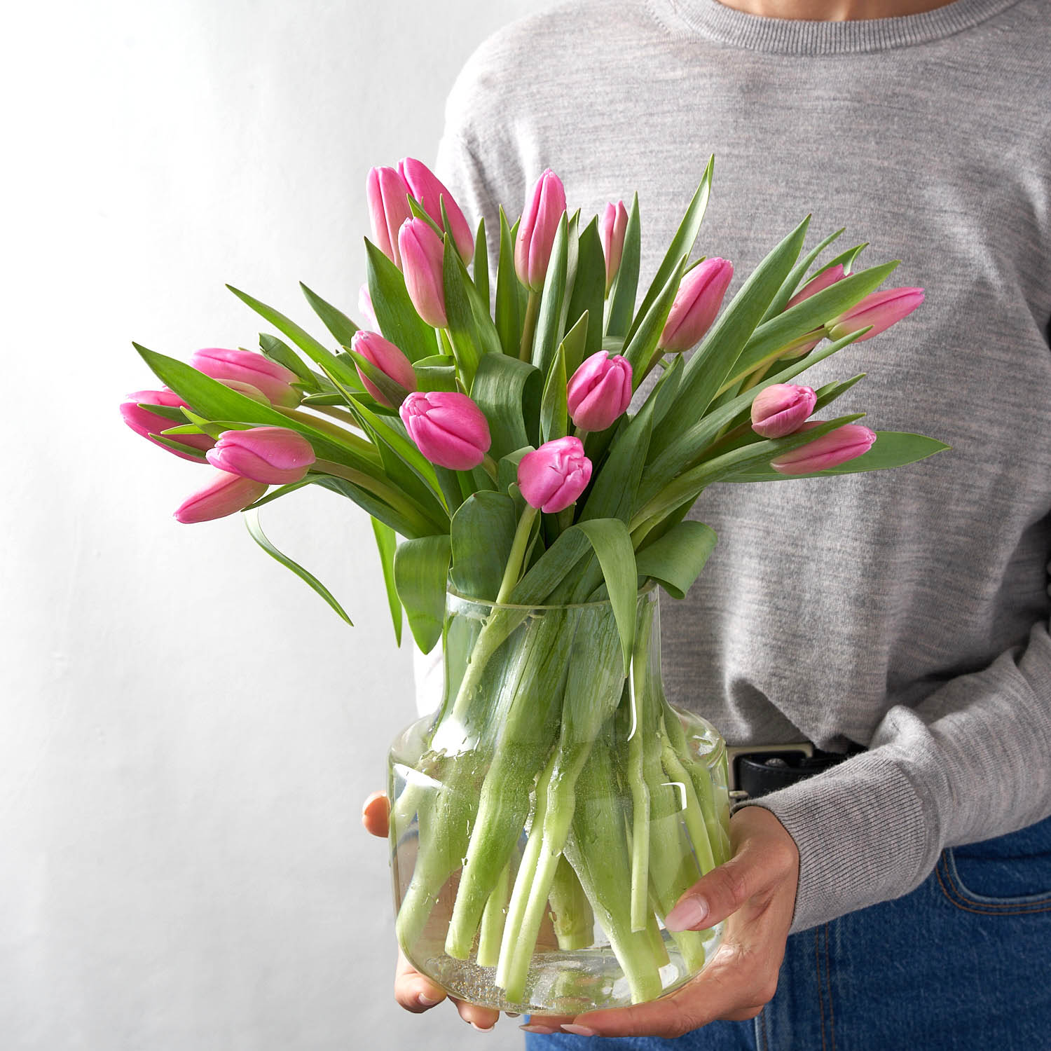 Woman in grey sweater holding a clear glass vase arranged with pink tulips