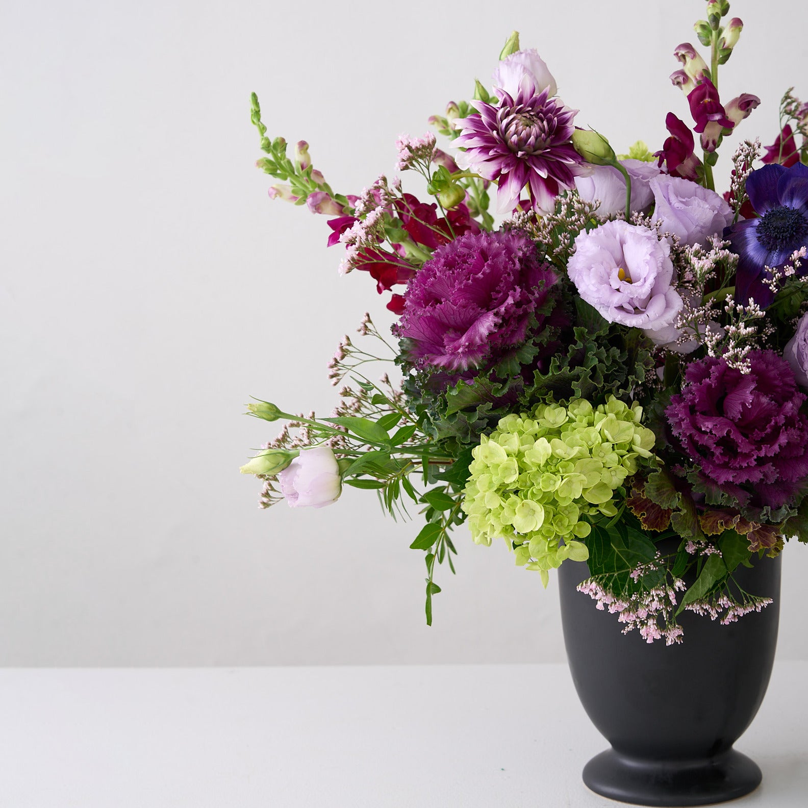 Halloween arrangement of dark purple and mauve dahlias, snapdragons, cabbage and lisianthus in a black vase
