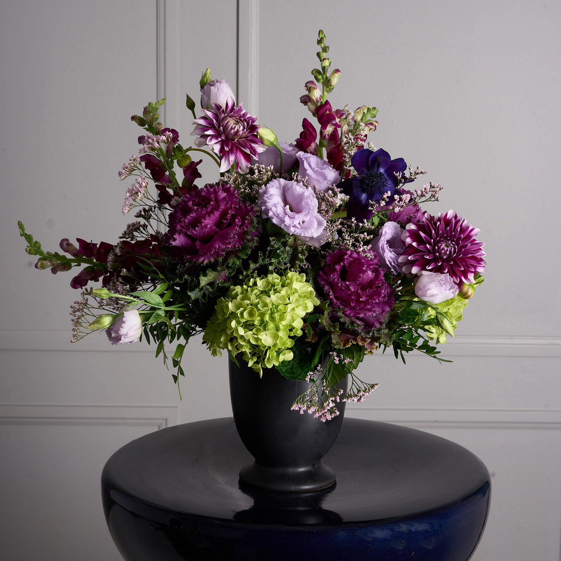 Halloween arrangement of dark purple and mauve dahlias, snapdragons, purple and green cabbage, and lisianthus in a black vase.