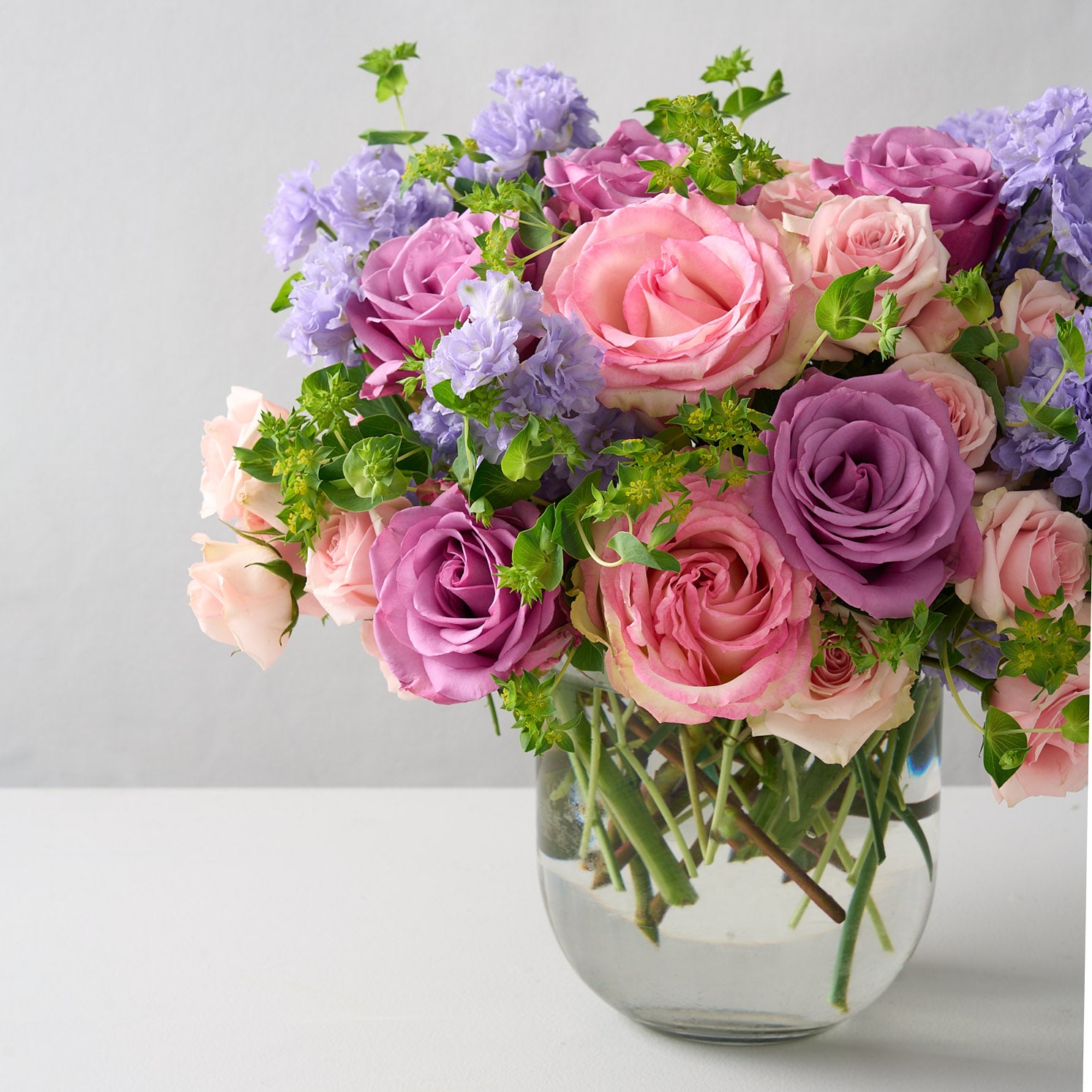 soft pink spray roses, green Bupleurum, lavender delphinium, Piacere roses, and Esperance Arranged in a round-bottom vase