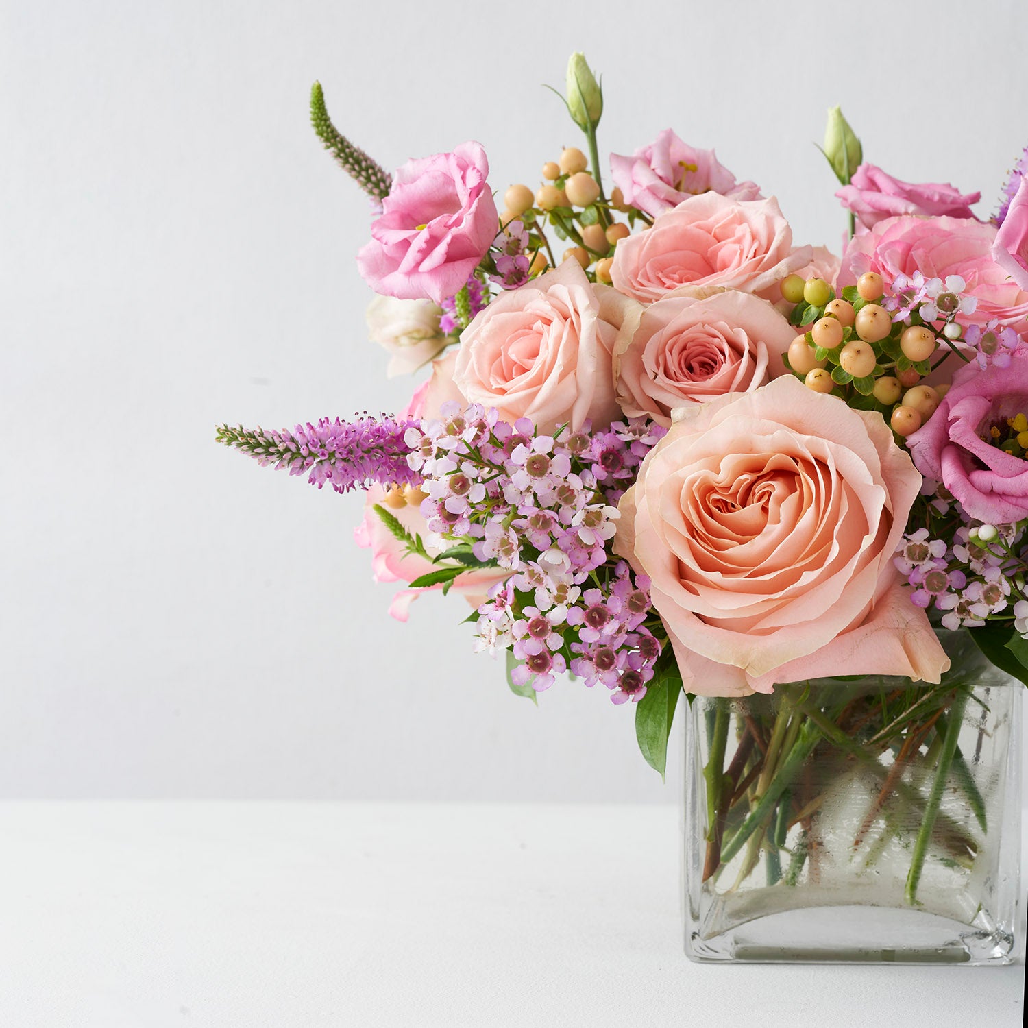 Arrangement of soft peachy roses, pink wax flower, pink lisianthus and pink veronica in glass vase on white background.