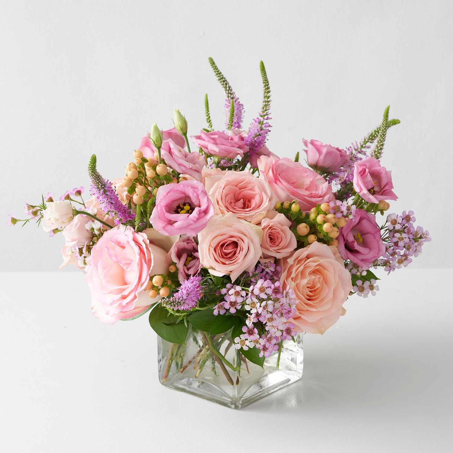 Arrangement of soft peachy roses, pink wax flower, pink lisianthus and pink veronica in glass cube centered on white background.