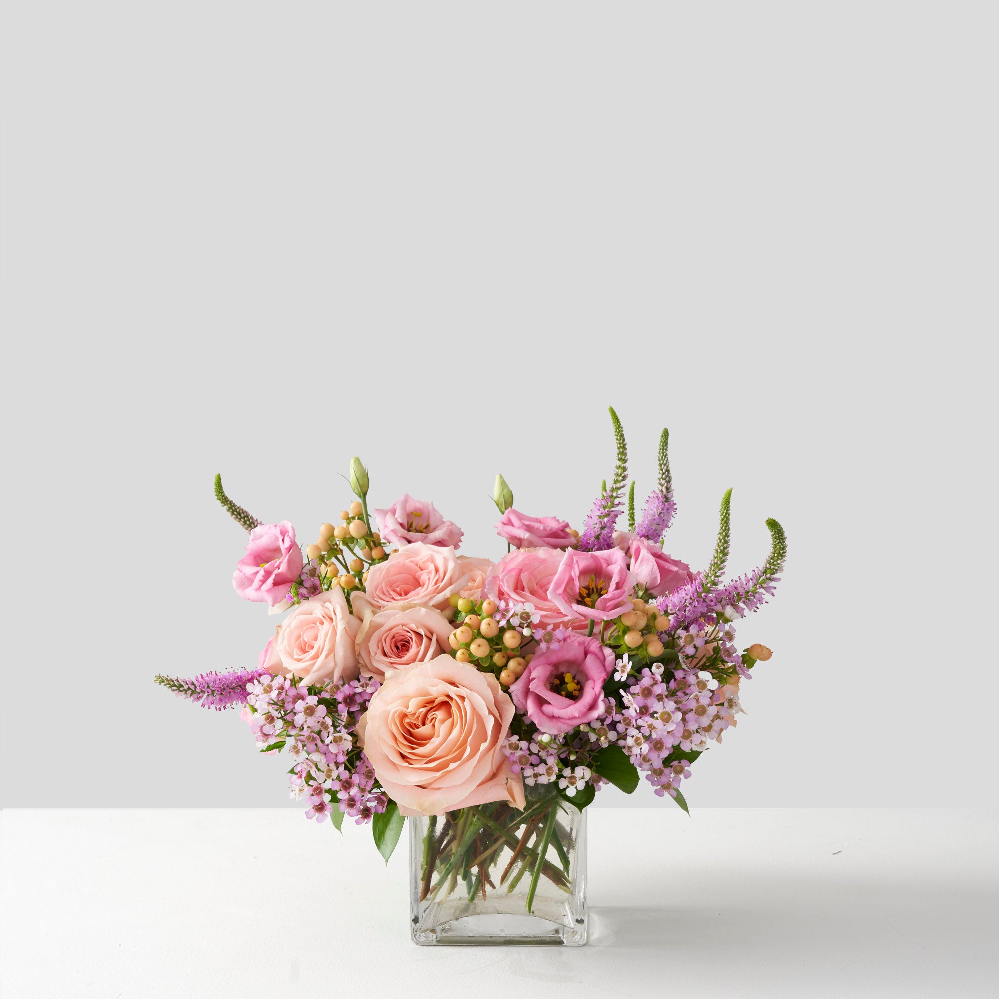 An arrangement of variouse pink and peach toned flowes, including roses, lisianthus, wax flower, and veronicain clear glass cube on white background.