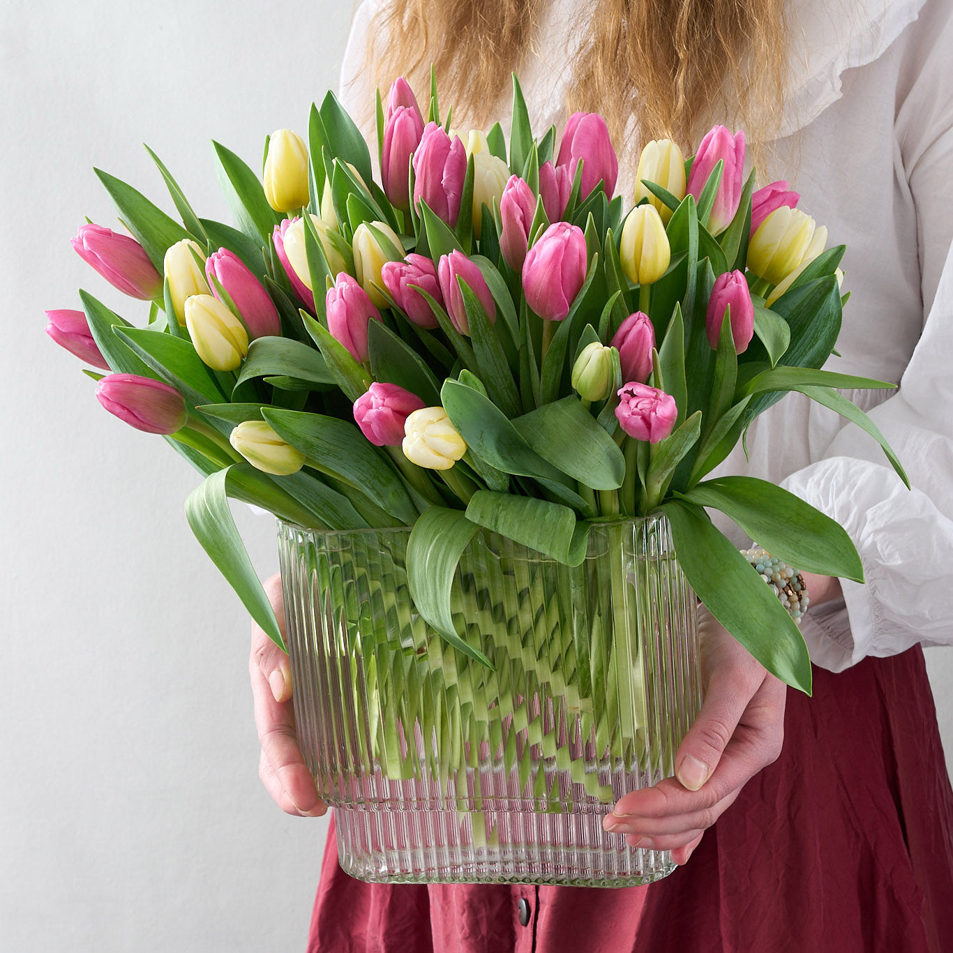Woman holding a floral arrangement featuring a mix of pink and white tulips in an clear, oblong ribbed vase.