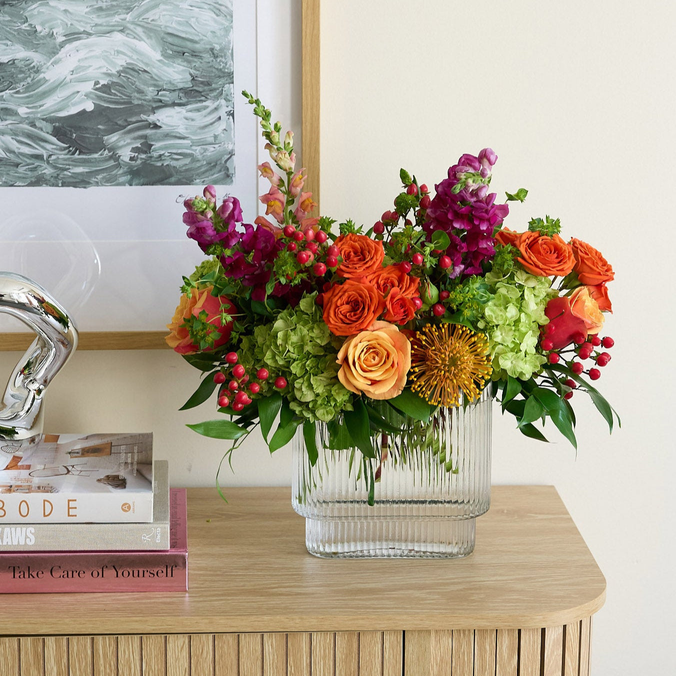 coral roses and hiypericum, green hydrangea , orange spray roses, fuchsia snapdragons, and protea in a clear square vase on credenza with books