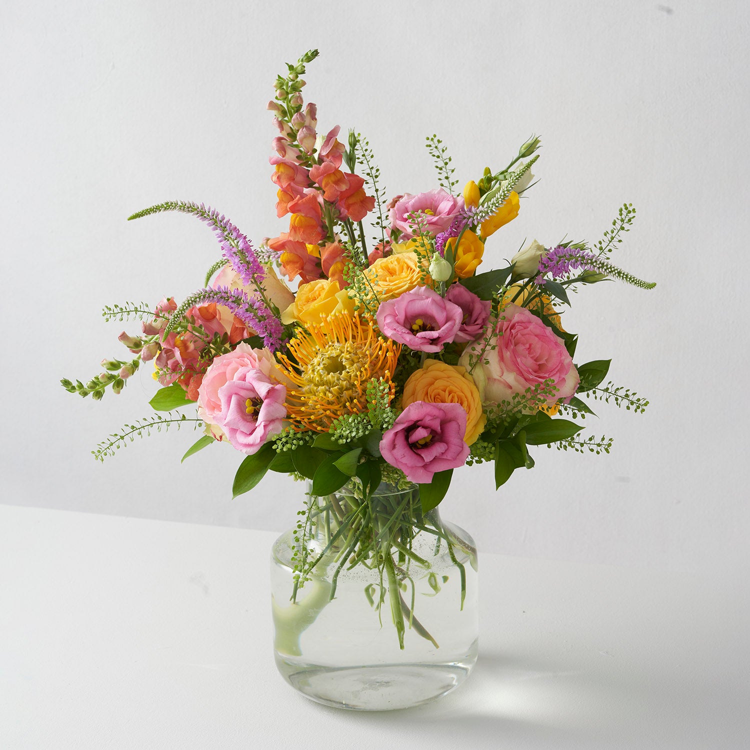 Arrangement of pink, yellow, and orange flowers in glass vase on white background.
