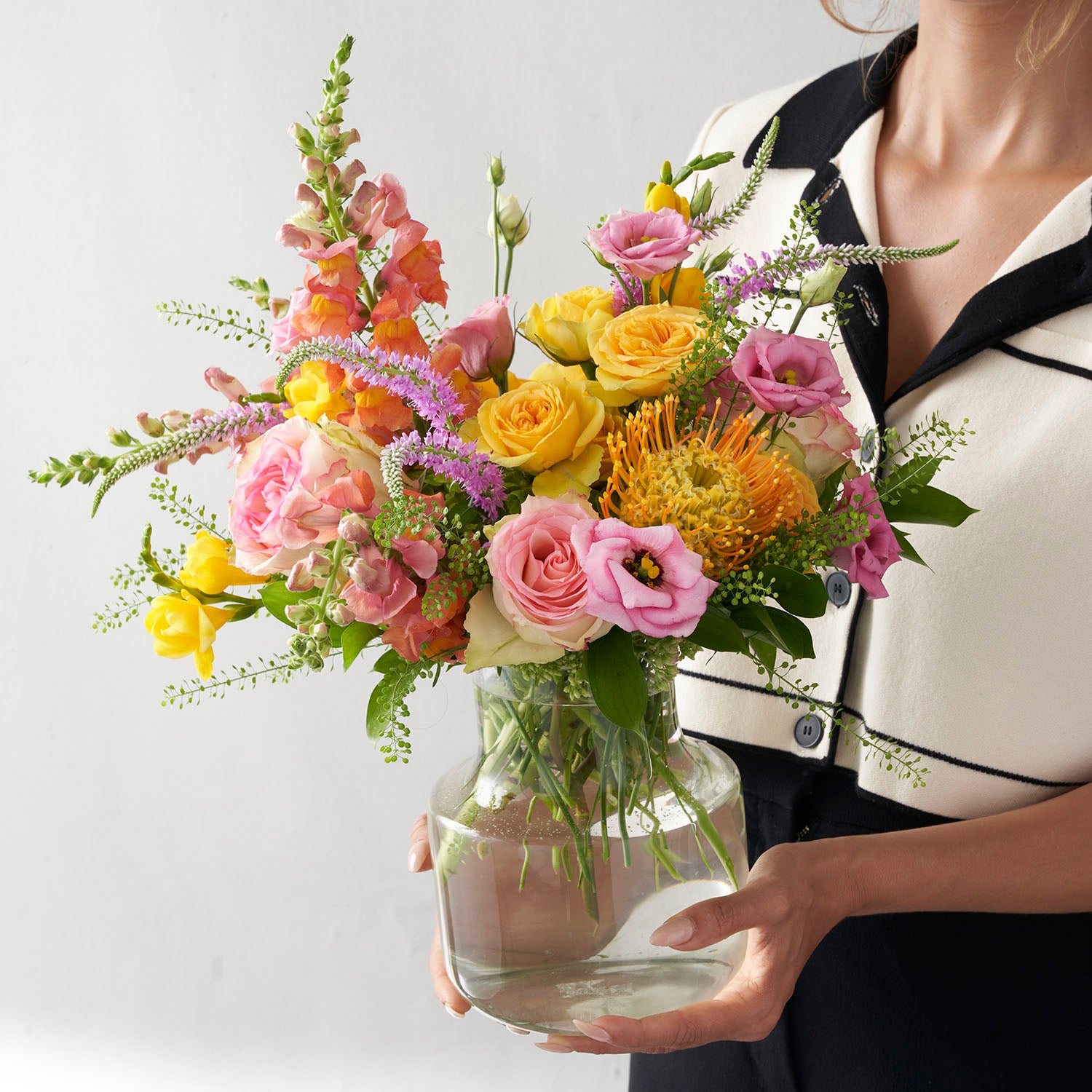 Woman in black and dress holding glass vase full of yellow, pink and oange flowers. 