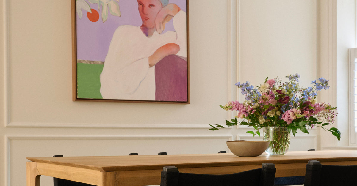 A framed art print depicting a stylized portrait of a woman, hanging on a cream-colored wall above a wooden dining table with a vase of colorful flowers.