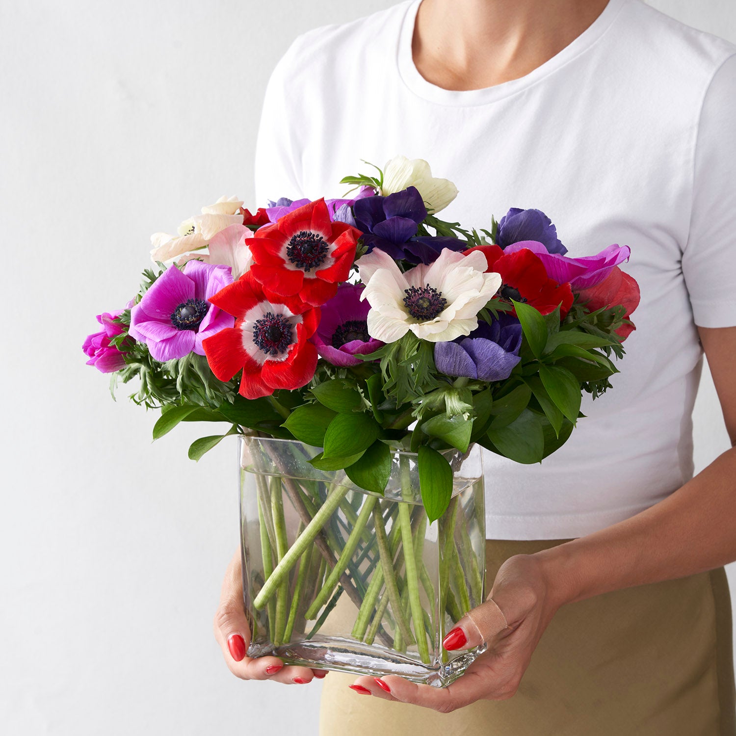 Woman in white shirt holding clear glass vase filled with multi coloured anemonie flowers.