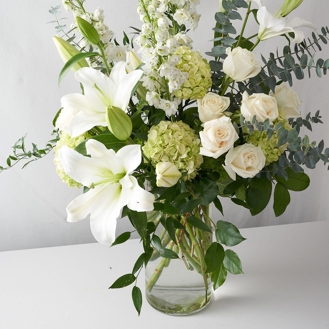 White lilies, white roses, green hydrangea and other greenery in a clear glass vase.