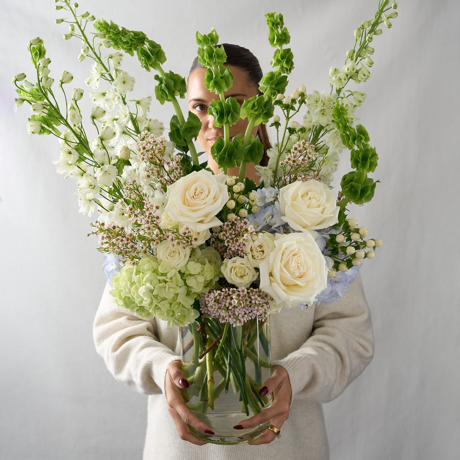woman holding a Tall flower arrangement with white delphinium, green bells of Ireland, blue & green hydrangea, cream candlelight roses