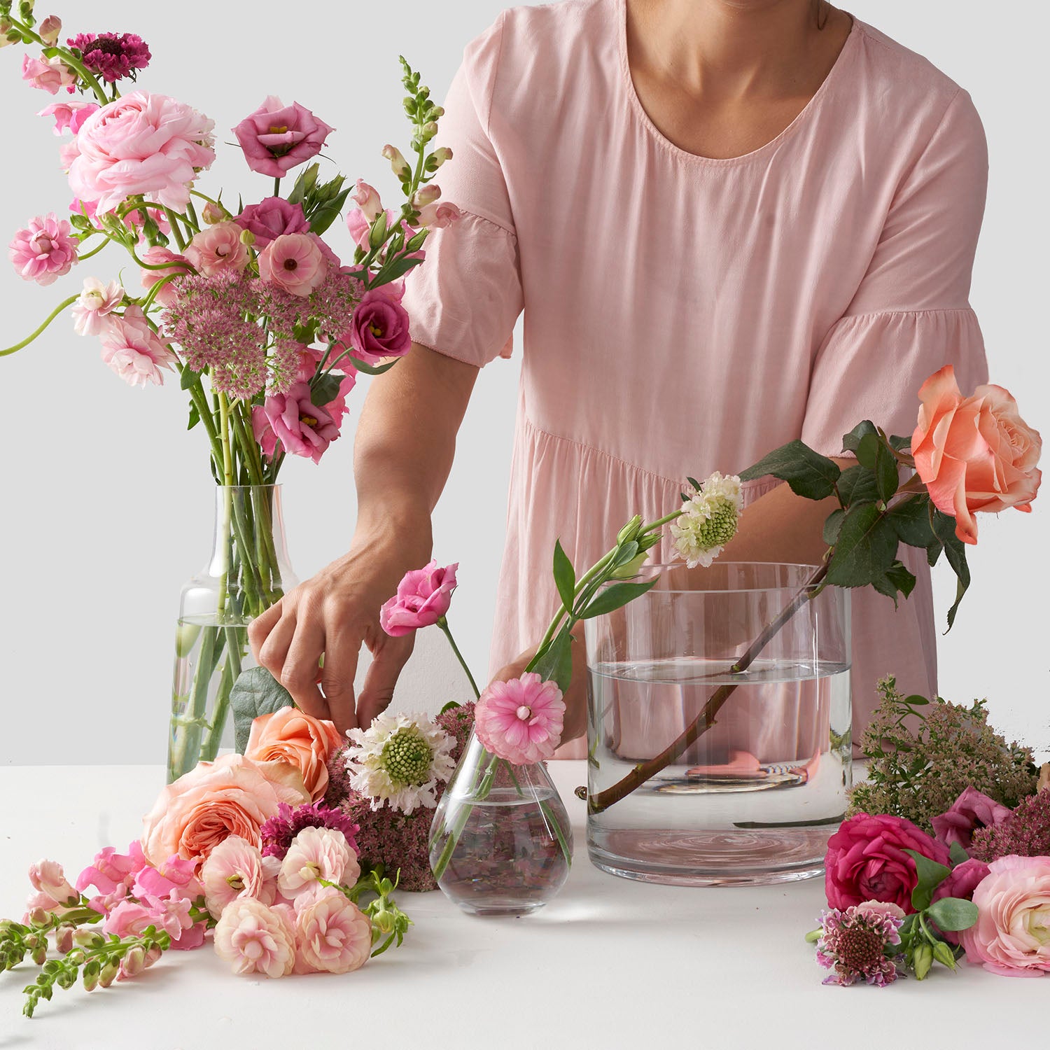 Woman in pink dress standing behind white table. Table holds clear three glass vases filled with water and bunches of pastel coloured flowers, including roses, lisianthus, ranunculus and snapdragons.