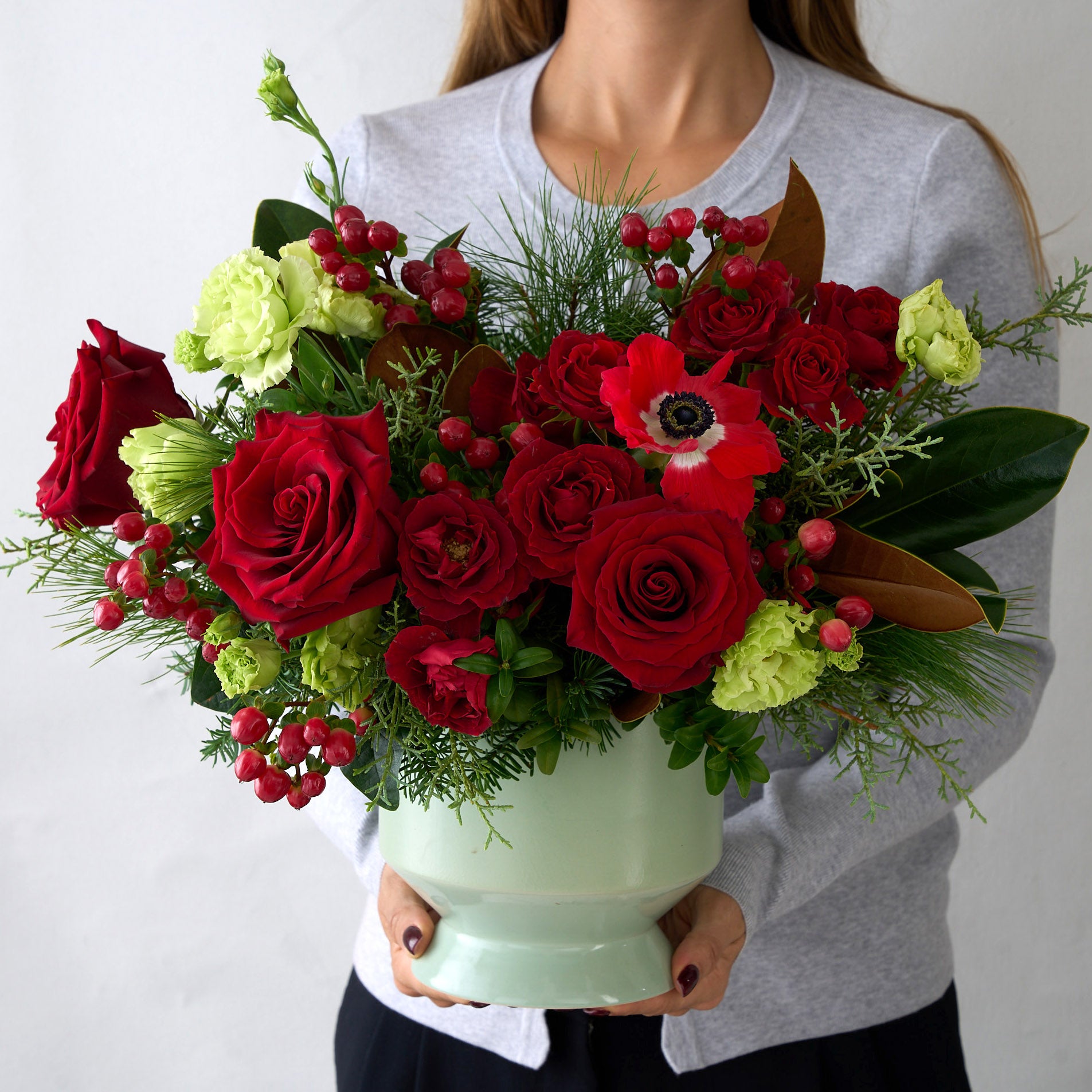 Woman in grey sweater holding a Christmas arrangement with double green Lisianthus, red roses, hypericum, and winter greens in a sage pedestal ceramic vase.