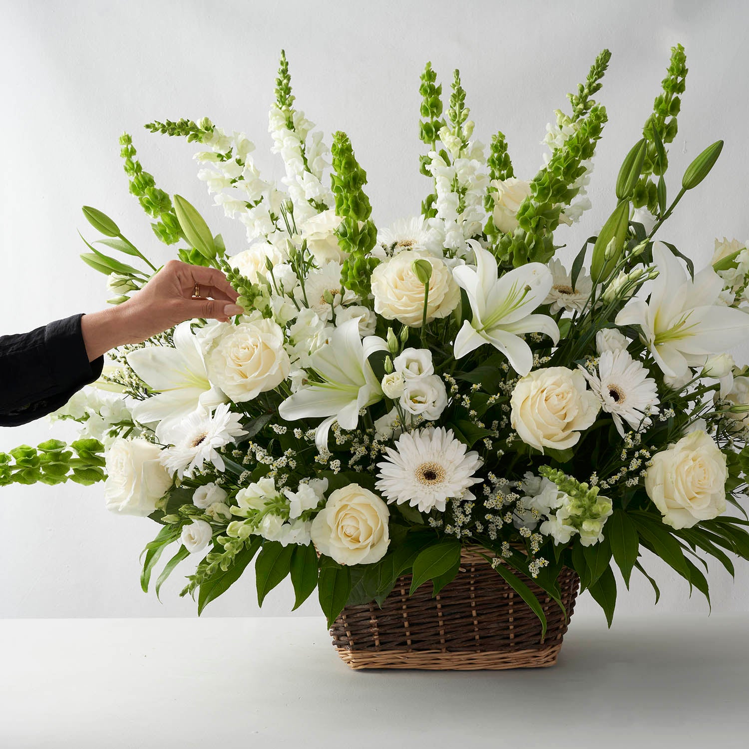 One hand adjusting flowers in an arrangement of all white flowers in a natural basket.