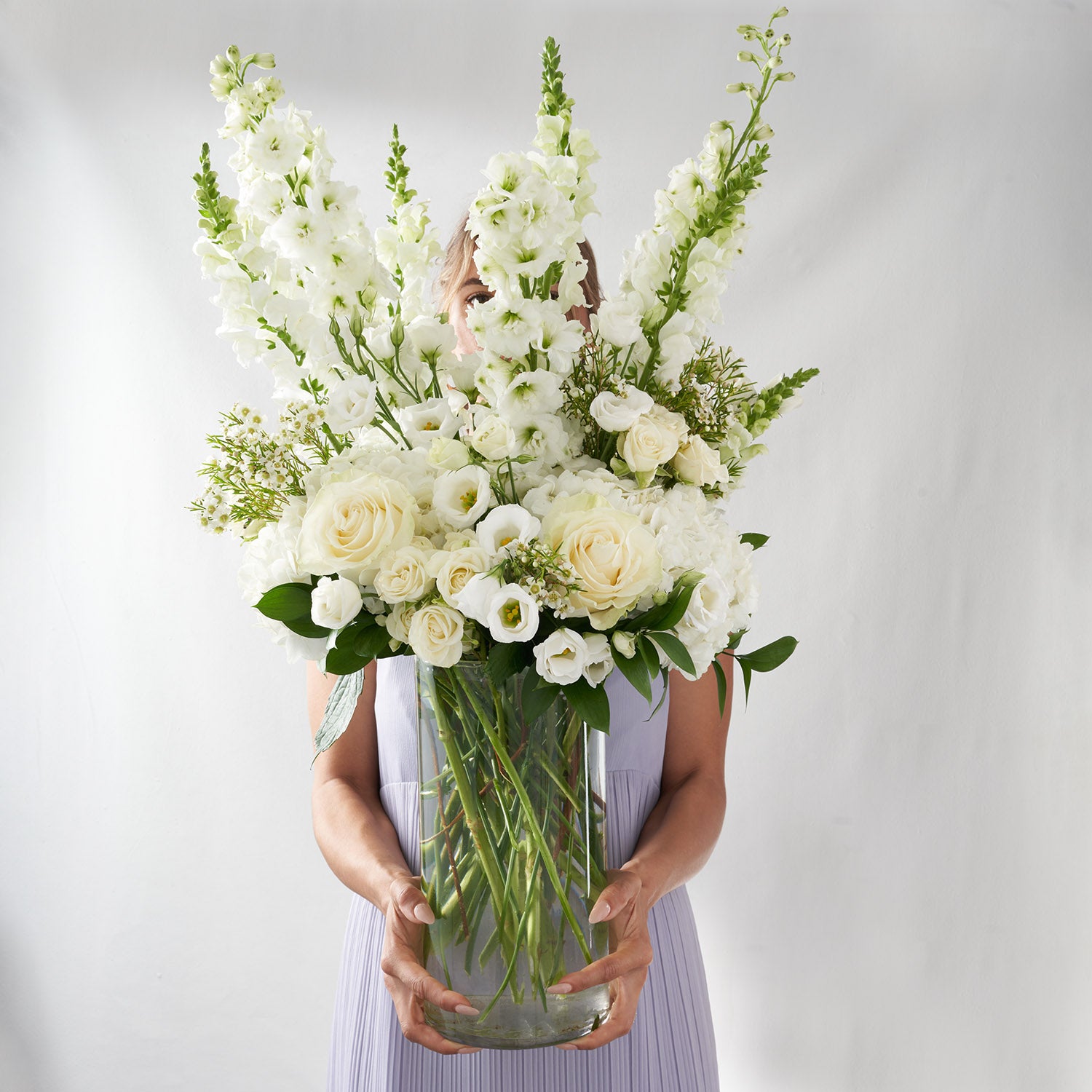 Woman in mauve dress holding large vase of white flowers including white roses, delphinium, and lisianthus.