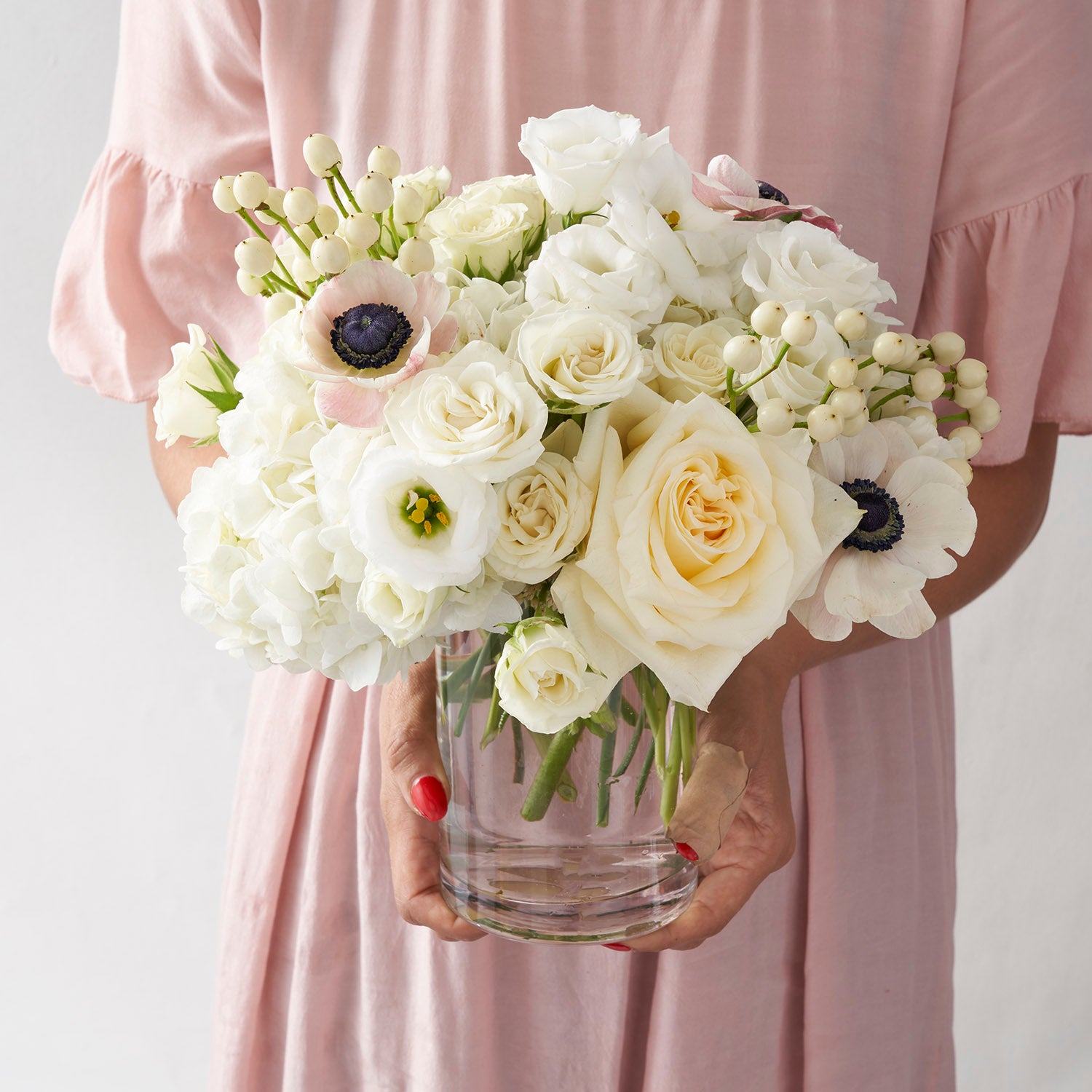 Woman in pink dress holding clear glass vase filled with cream and white flowers.