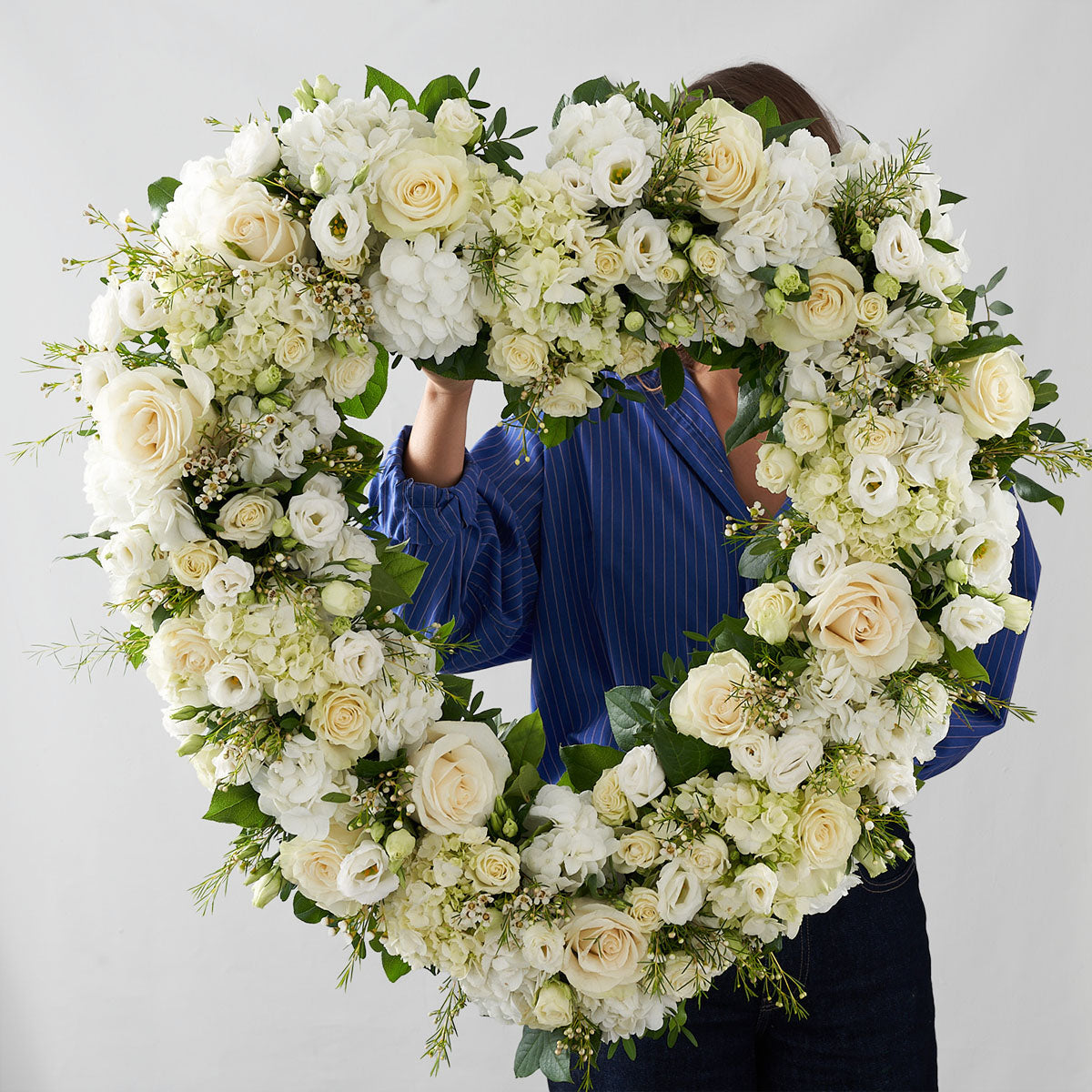 Woman in blue arranging a heart-shaped wreath adorned with white roses, white hydrangea, white lilies, white anemones and other greenery