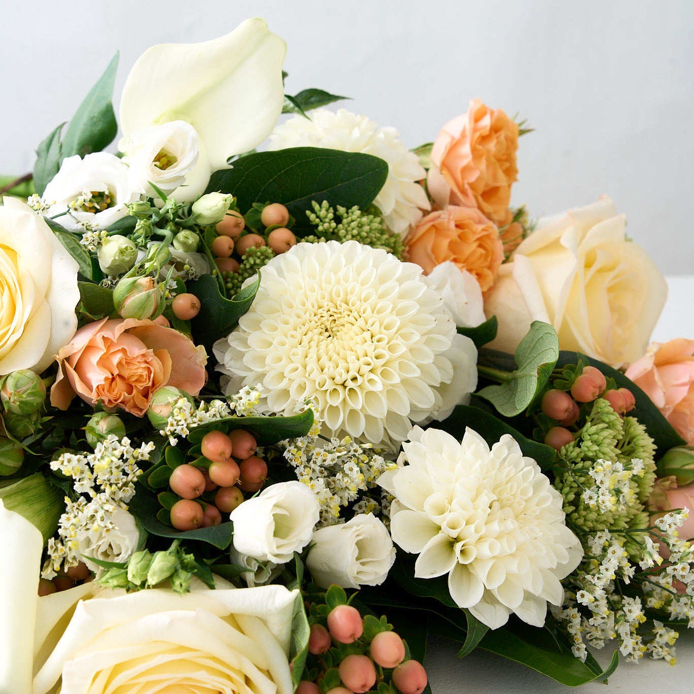 close up of a bouquet of white pompom dahlias and Lisianthus, peach hypericum and rose with greens on a light background