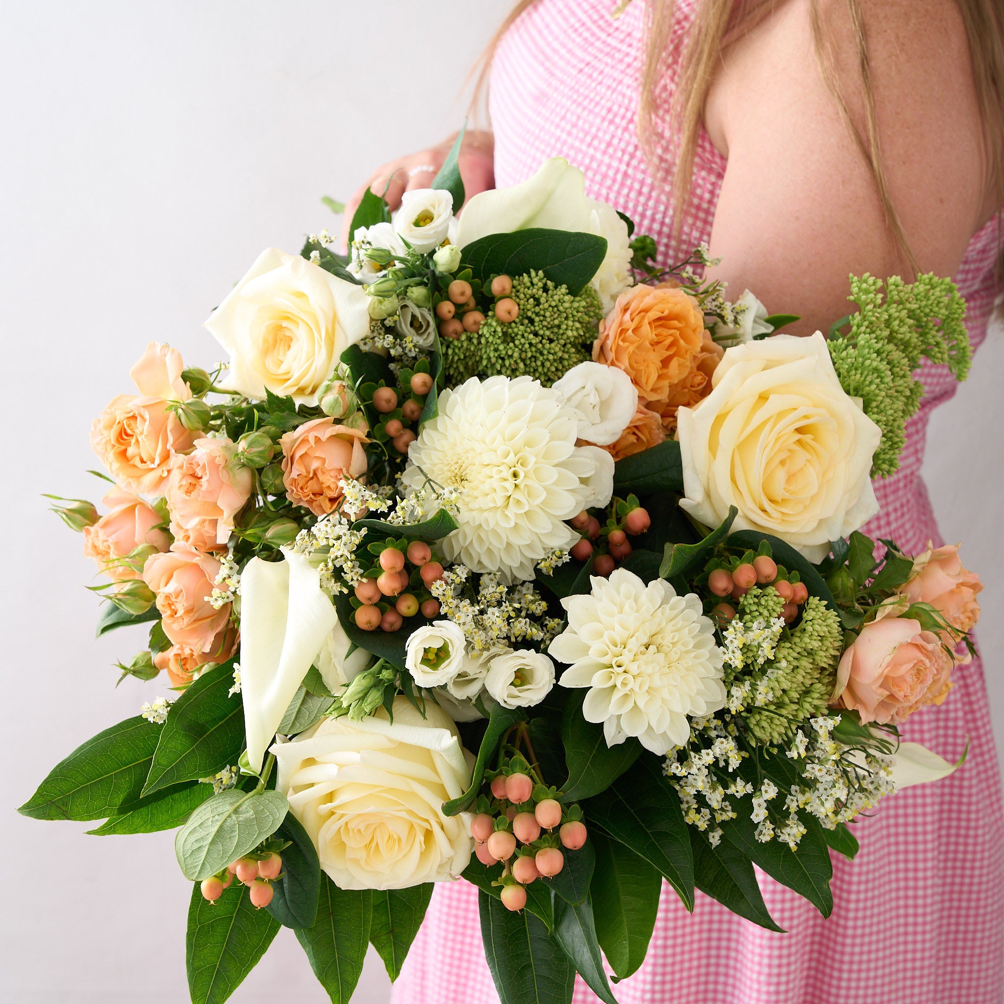 Bouquet of white dahlias, candle light roses, peach roses and greens held by a woman wearing a red gingham summer checkered dress.