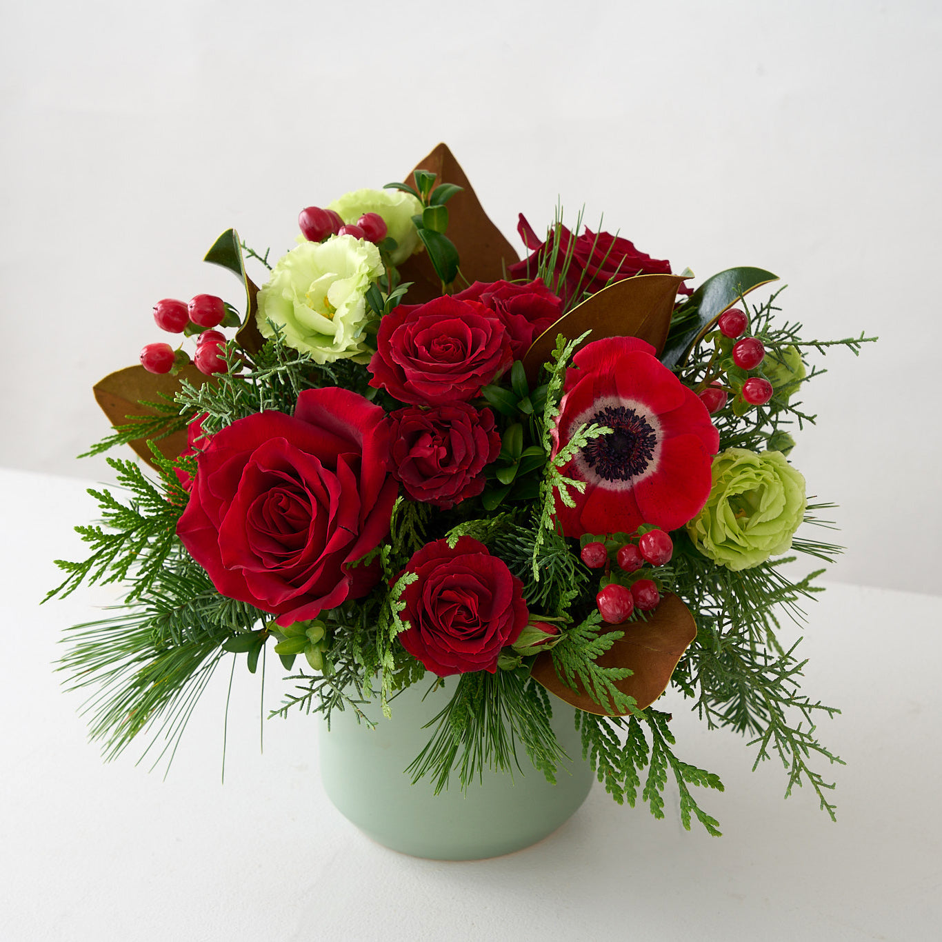 Christmas arrangement of red roses, hypericum, anemones, magnolia leaves, green Lisianthus and winter greens in a small sage vase