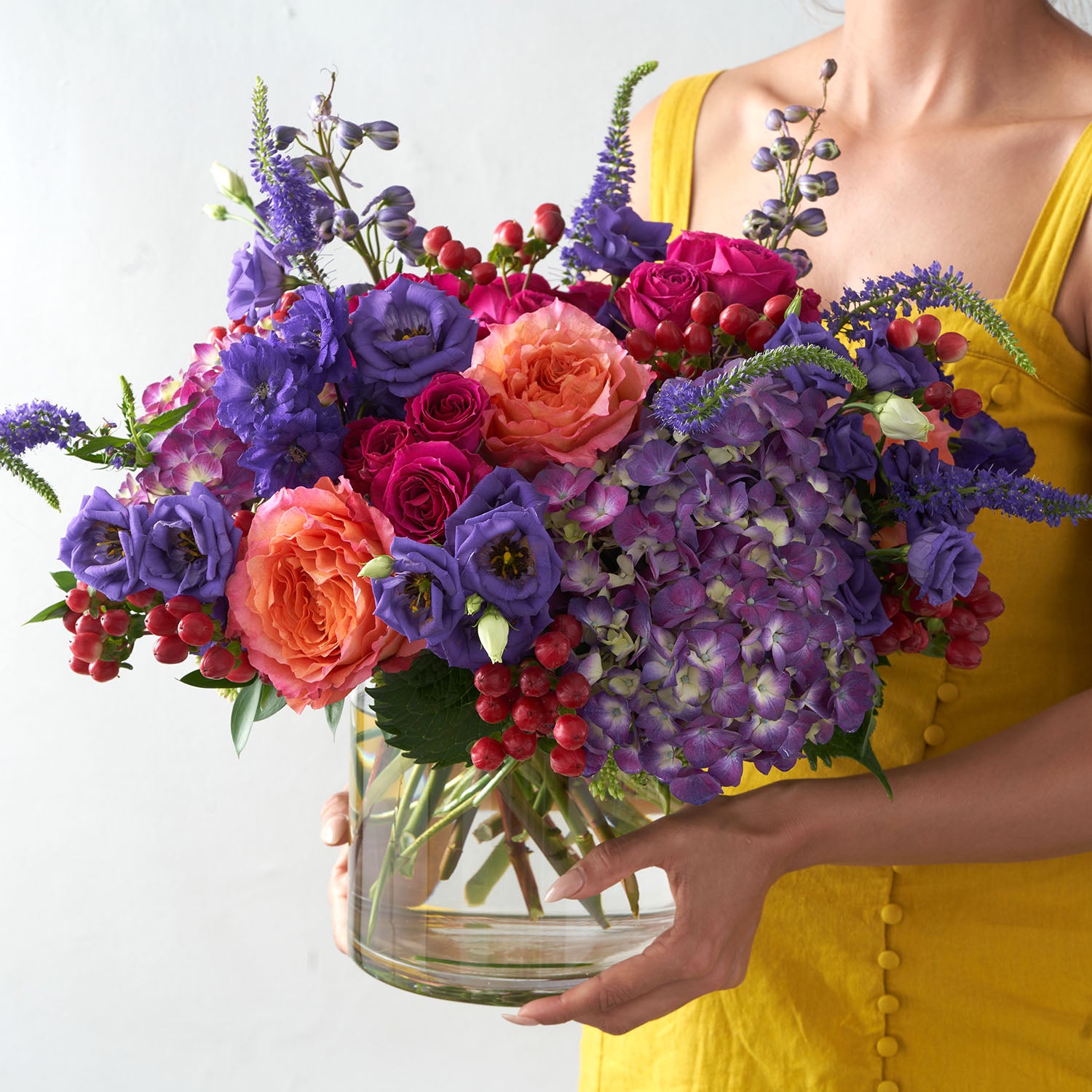 Woman in yellow dress holding glas vase filled with deep purple, orange, and fuchsia flowers.