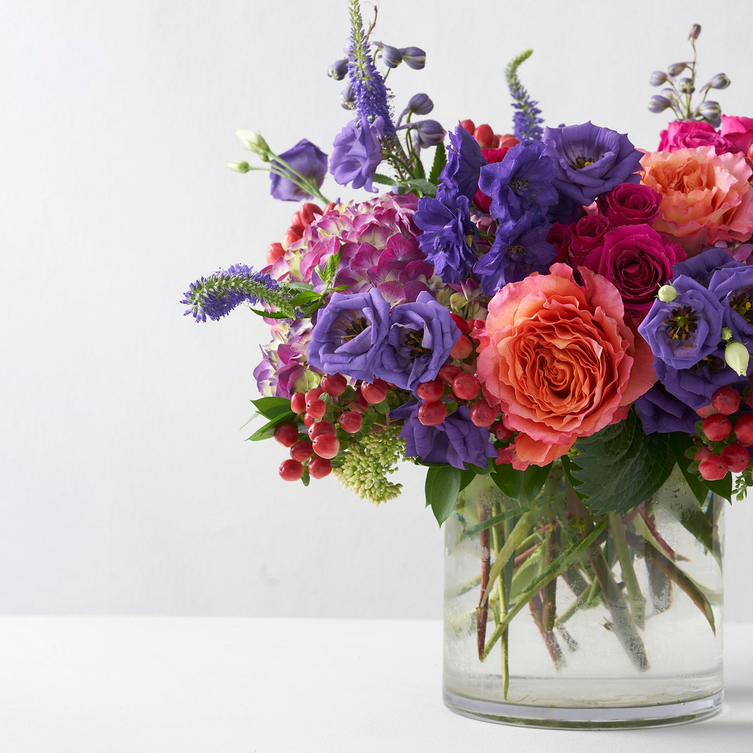 Arrangement of orange, purple, and hot pink flowers arranged in glass vase on white background.