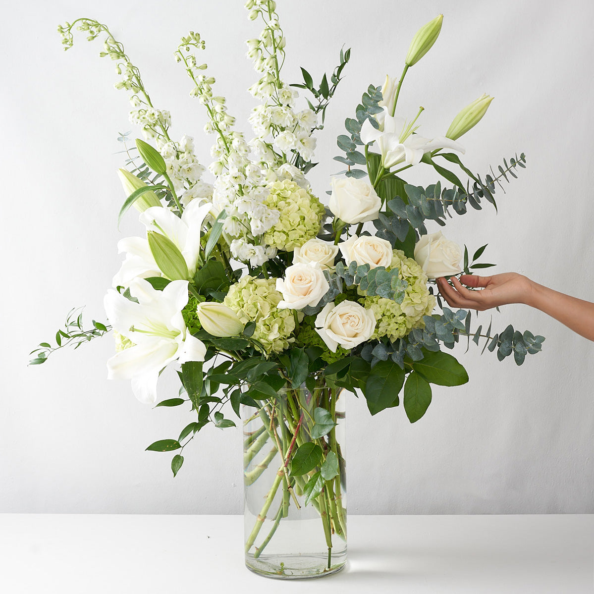 A clear glass vase containing, white roses, green hydrangea, white lilies and lots of greenery