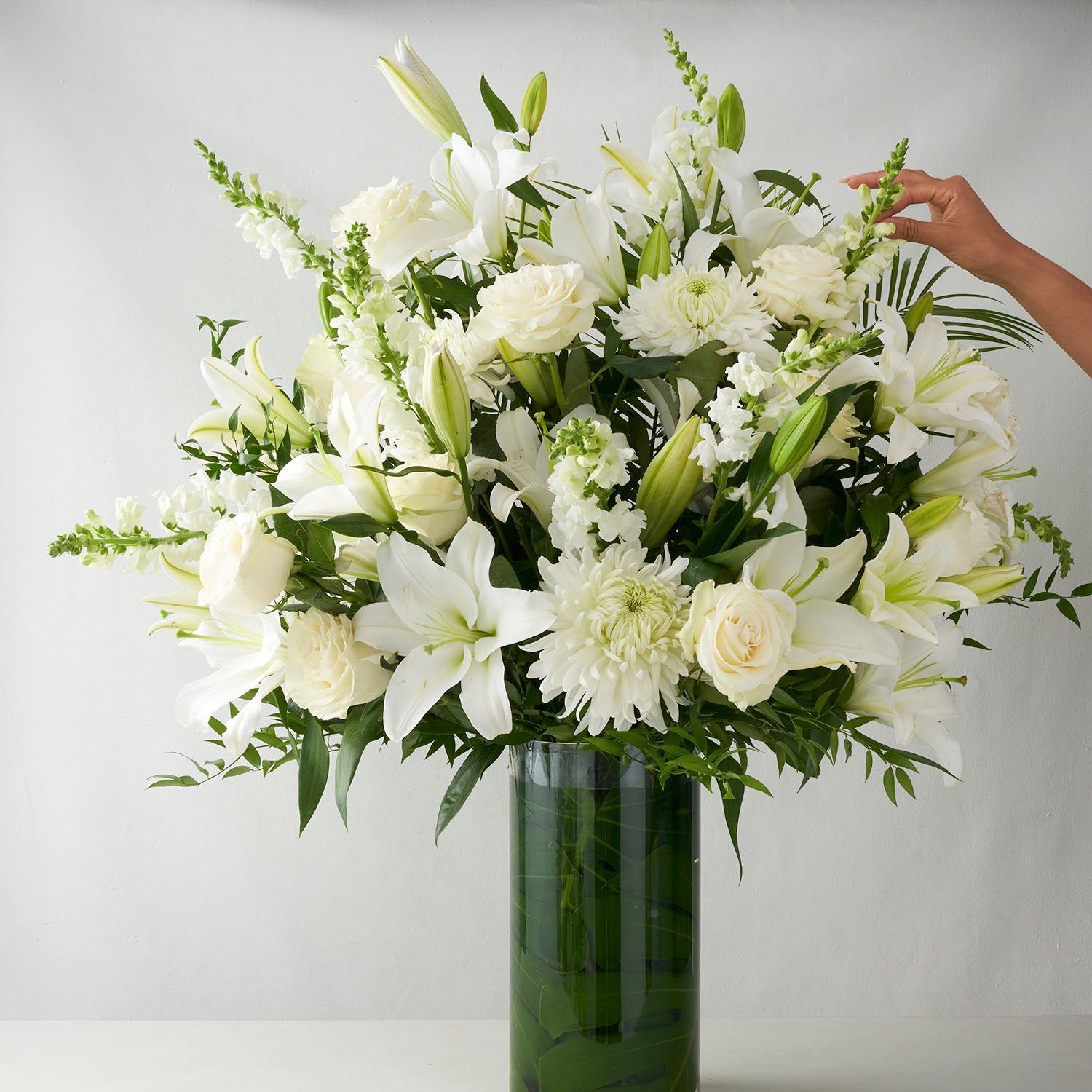 One hand adjusting flower in large glass vase full of white flowers.