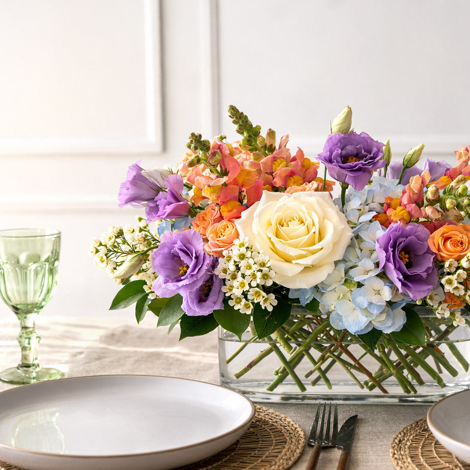 Colorful Easter floral centerpiece arrangement in a glass vase on a table with plates and cutlery.