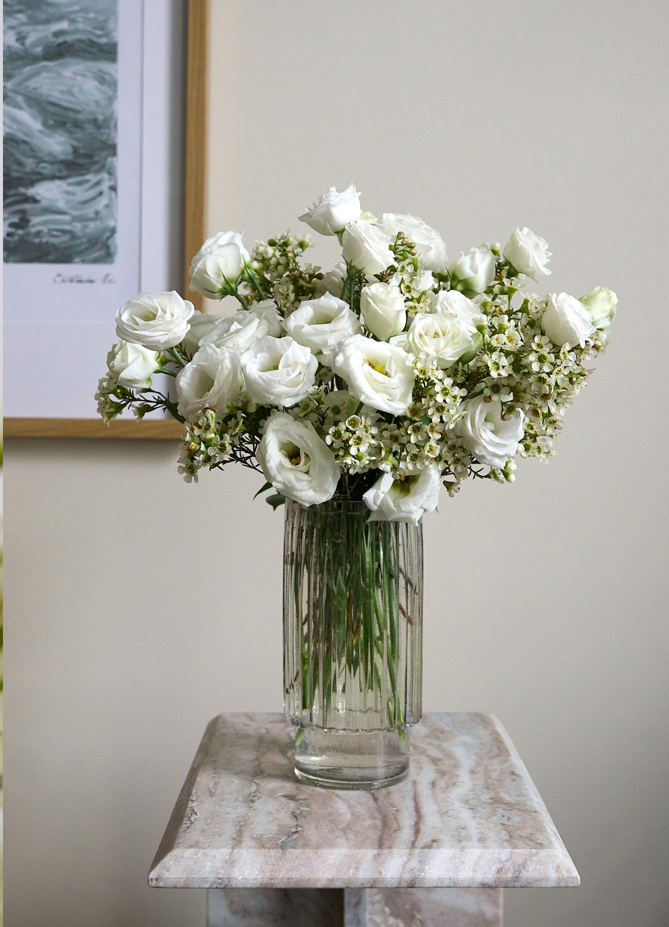 White lisianthus and wax flowers in a tall beveled glass vase on a side table