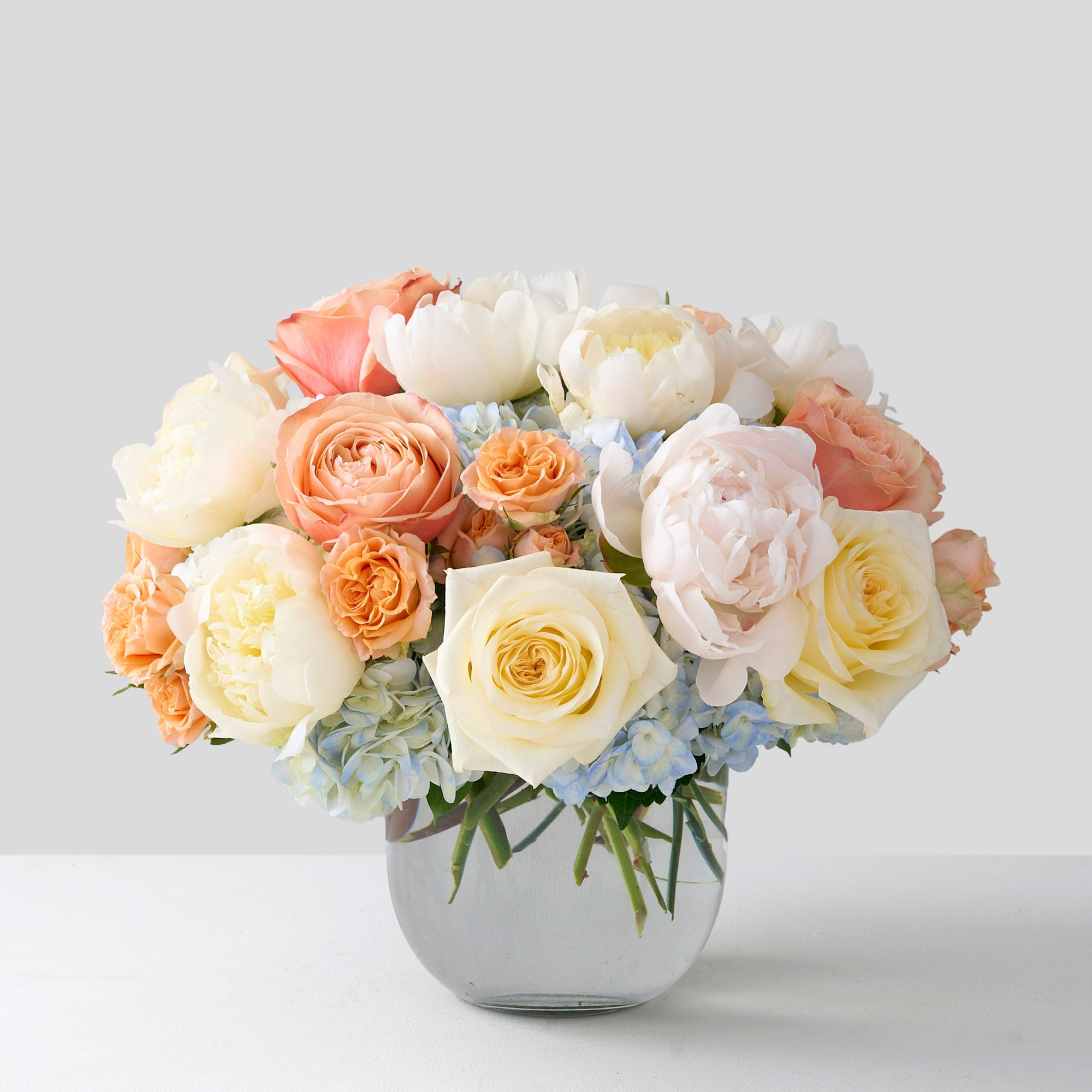 A glass vase filled with a bouquet of white and peach roses, cream peonies, and light blue hydrangeas, set against a plain grey background.