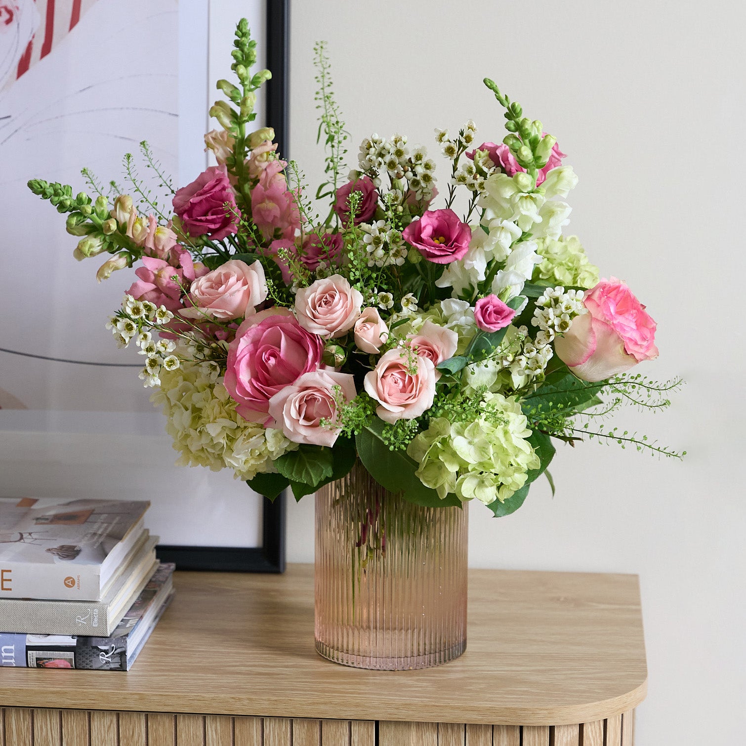 tall pink gold ribbed vase with pink and white flowers on a credenza with books and picture in the background