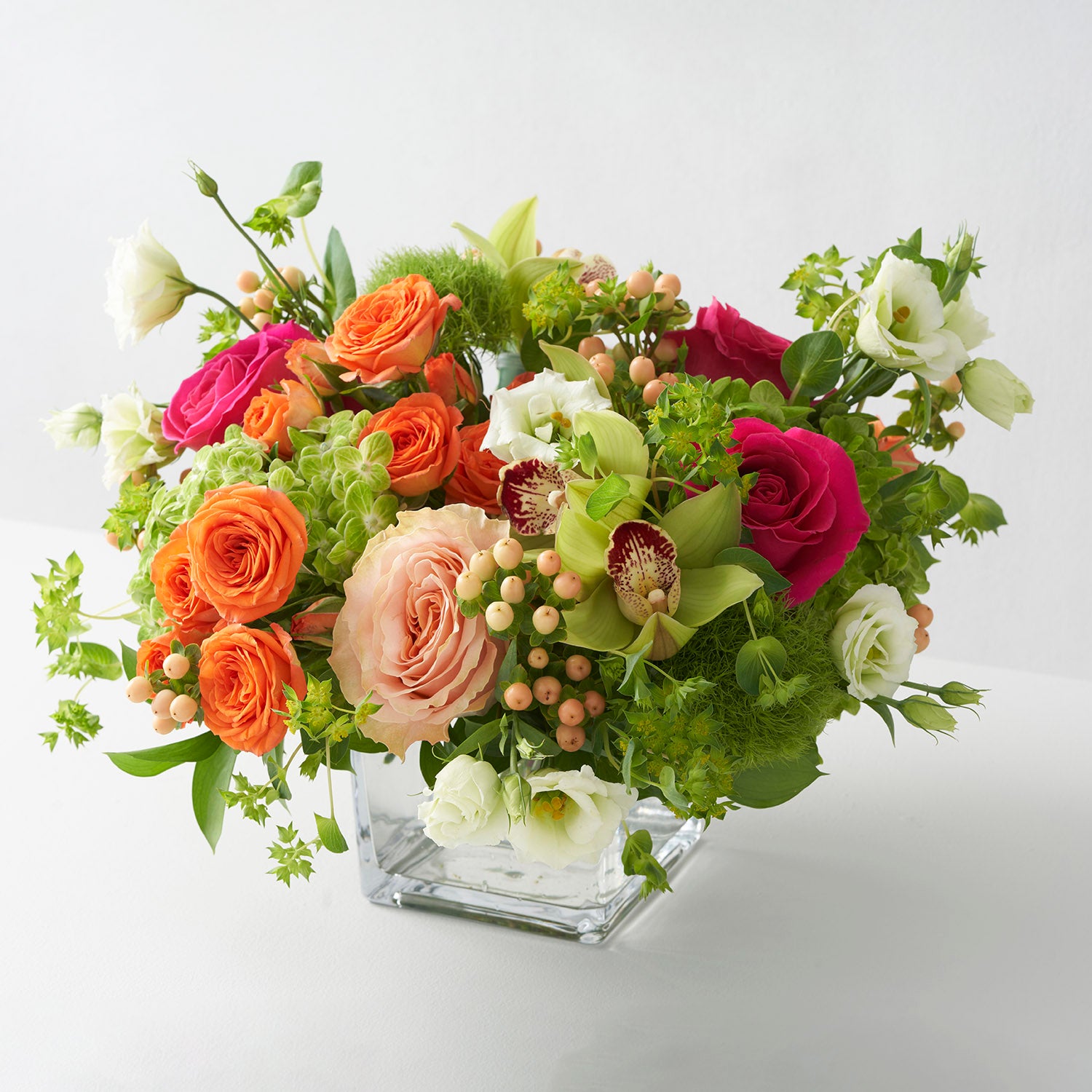 Orange, peach, pink, and green flower arrangement in simple glass vase on white background.