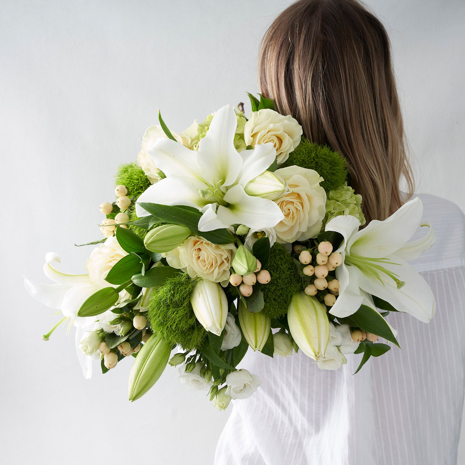 Woman in white shirt holding bouquet of white roses, white lilies, cream berries, green hydrangea and green dianthus.