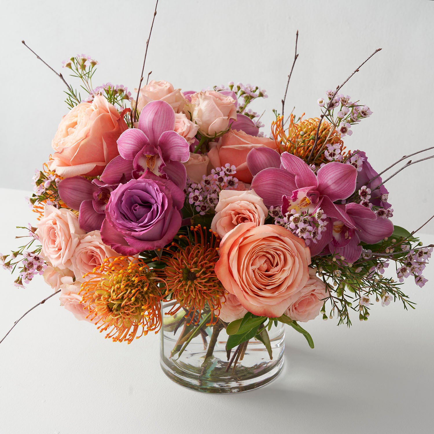 Peach and plum coloured roses with gold protea and plum cymbidium orchids, arranged in a glass vase on white background.