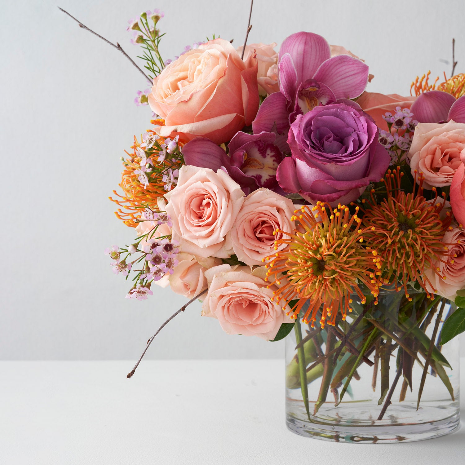 Roses, protea and cymbidium orchids arranged in clear glass vase off center of white background.