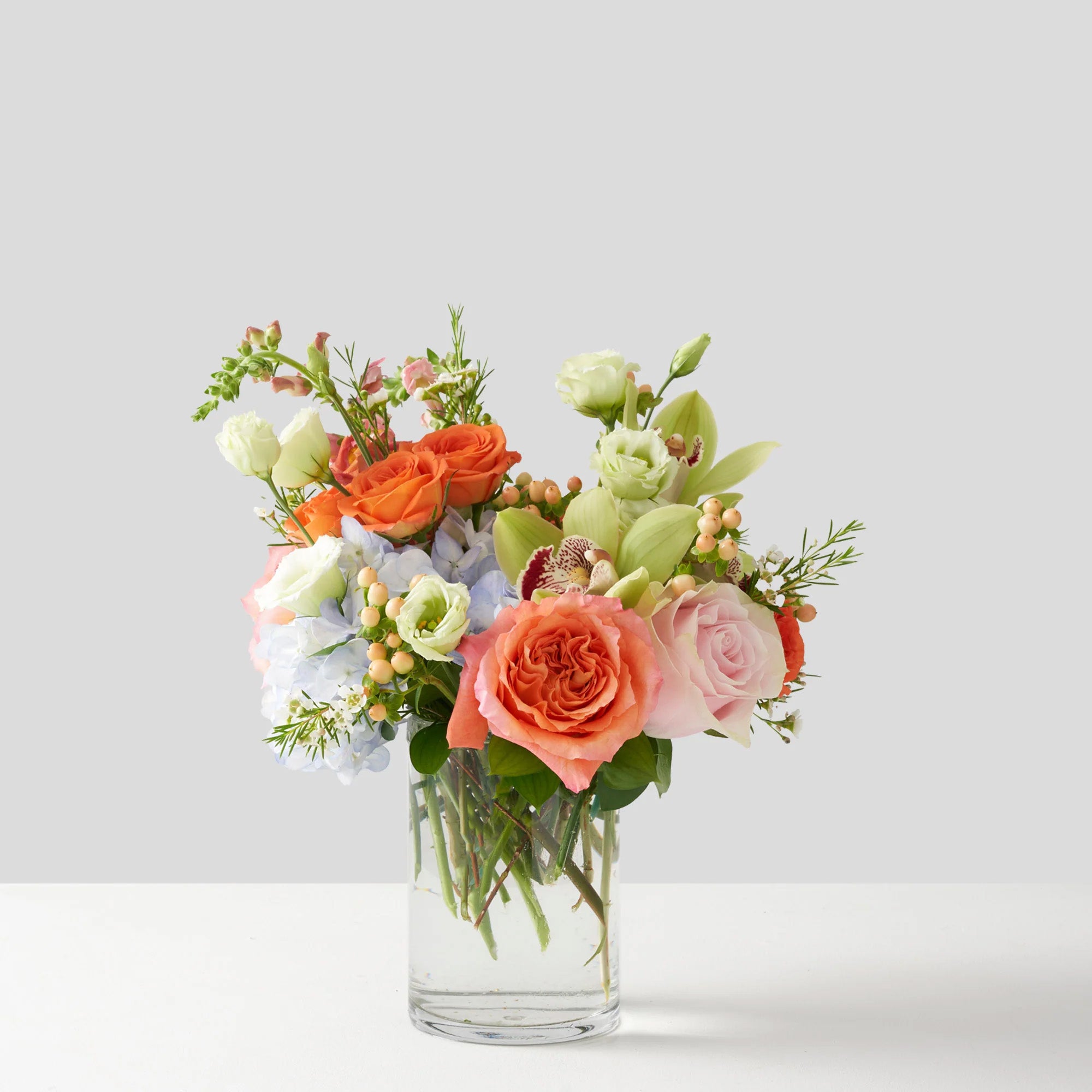 Clear glass vase on table with soft pastel coloured flowers, including orchids, orange roses, hydrangea and lisianthus